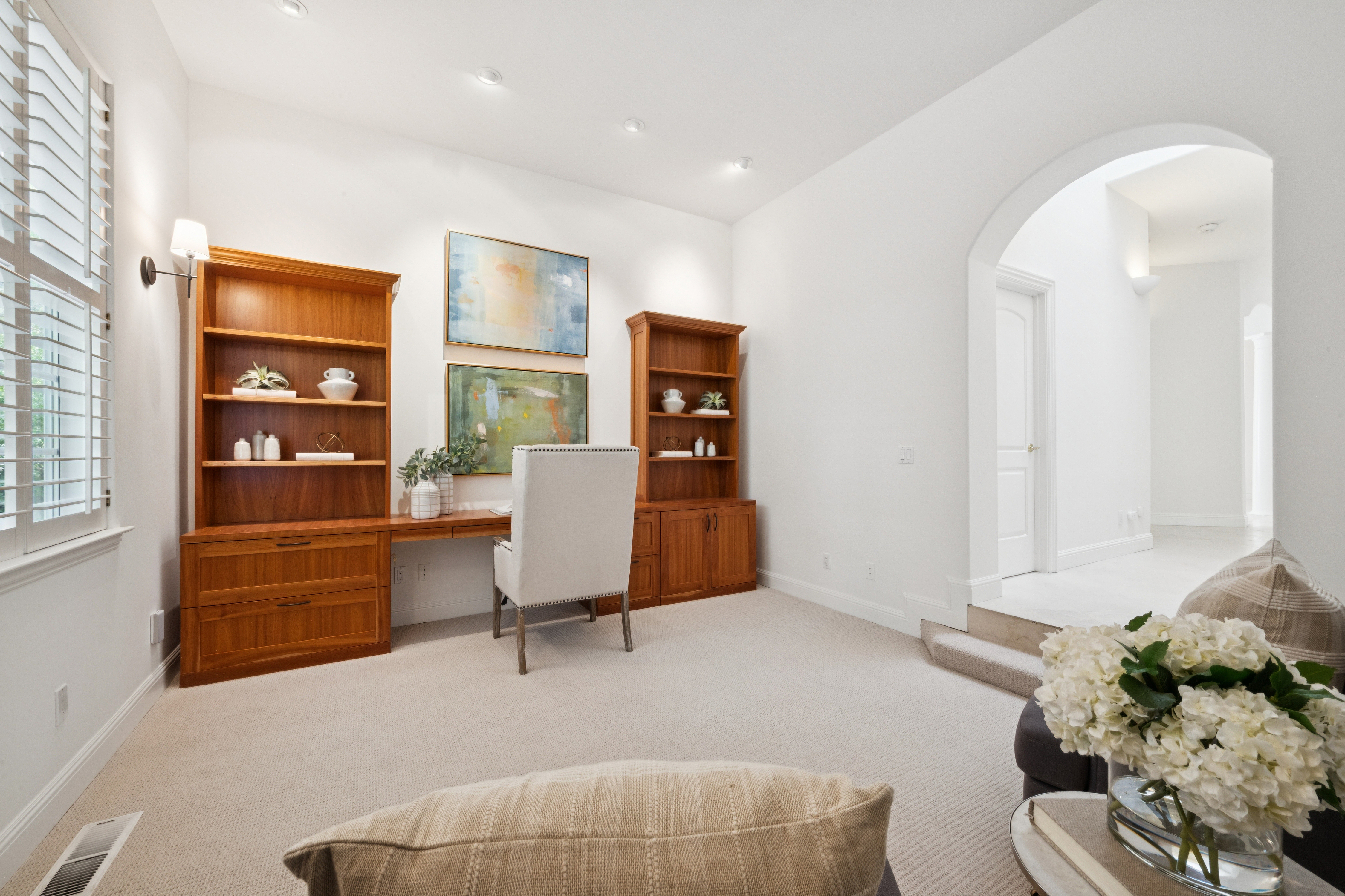 This sophisticated home office features custom built-in wooden shelving surrounding a built-in desk, providing a clean and organized workspace. A neutral-toned upholstered chair complements the light-colored walls and plush carpeting, creating a bright and professional atmosphere. The room also includes elegant wall sconce lighting and leads into a transitionary hallway area via a graceful arched entryway.