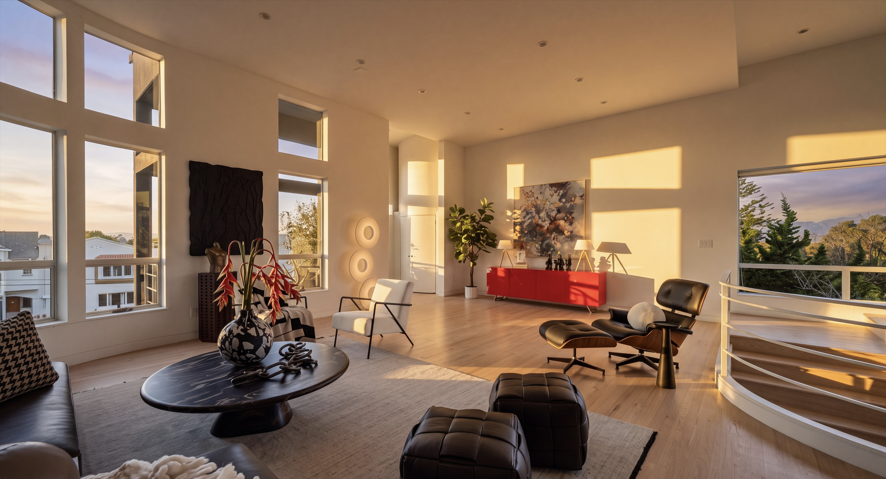 This spacious, light-filled living room features high ceilings and large windows that provide expansive views of the outdoors. The room is furnished with a modern black oval coffee table, a classic lounge chair with an ottoman, and two leather ottomans, all arranged on a neutral area rug over light wood flooring. A vibrant red console table adds a bold pop of color against the white walls, while a curved staircase railing on the right leads to an adjacent level.