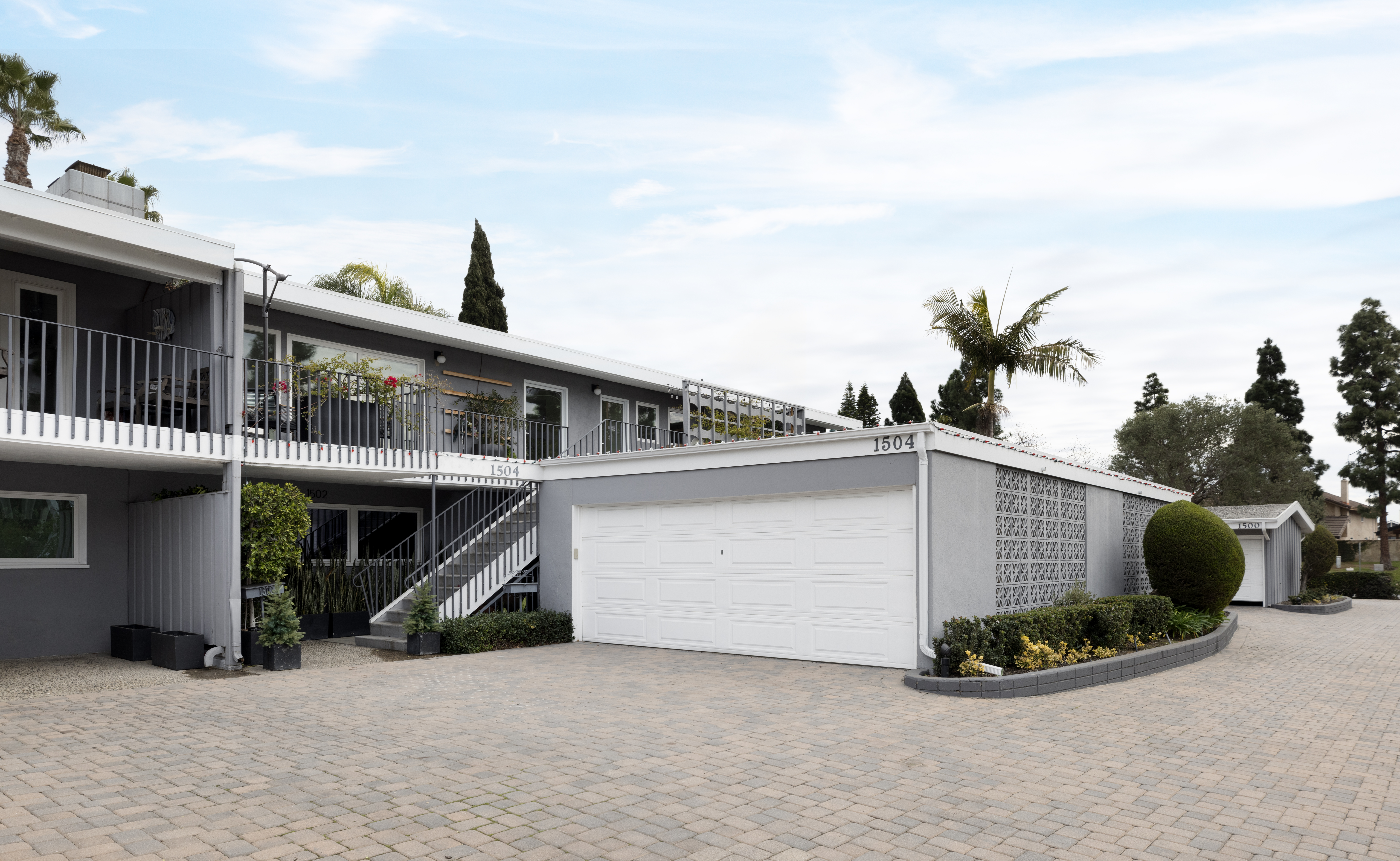 This mid-century modern apartment complex features a two-story gray facade with a prominent white garage door and a paved driveway. The building includes an upper-level balcony with metal railings and an exterior staircase leading to the second floor, accented by decorative breeze block walls. The scene is captured from a low-angle perspective, highlighting the clean lines of the architecture and the well-maintained landscaping with palm trees and manicured shrubs.