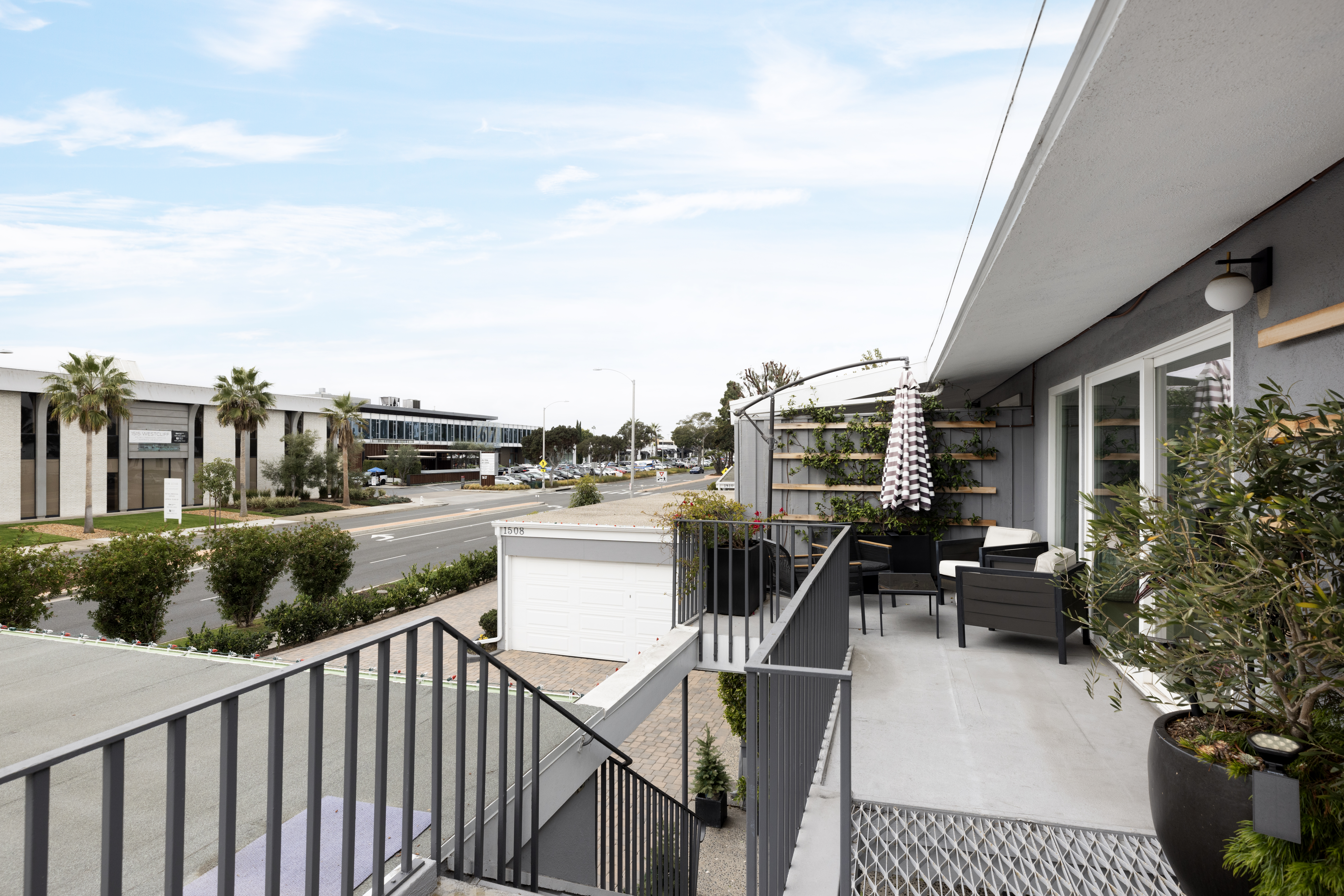 This elevated balcony offers a modern outdoor living space with dark-toned patio furniture and a striped umbrella, perfect for relaxing while overlooking the street below. The area features a metal railing, a decorative trellis with climbing greenery, and a large potted plant, creating a private and stylish retreat. The perspective captures the balcony's connection to the building's exterior and the surrounding neighborhood, emphasizing its functional design and urban setting.