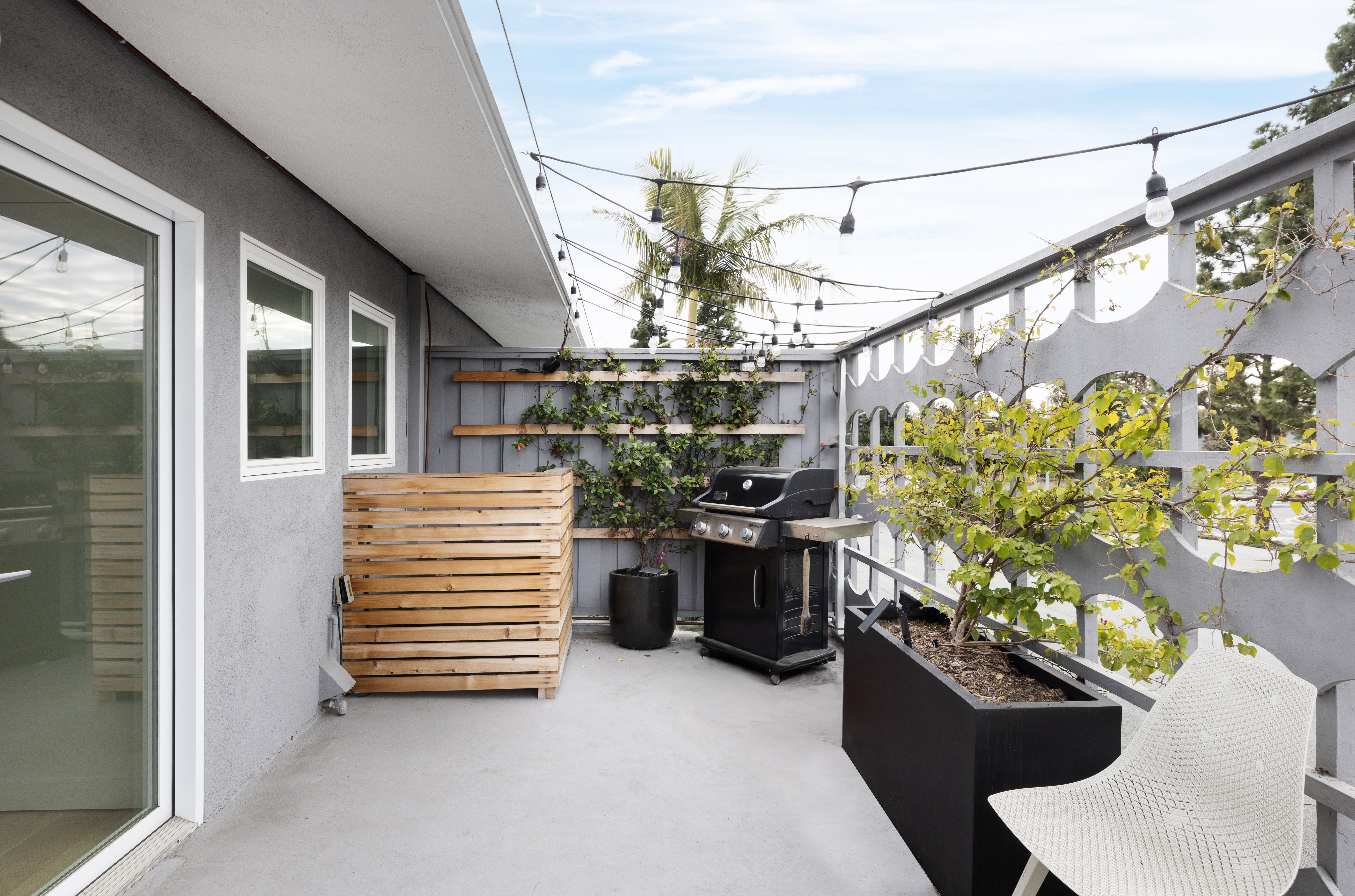 This modern balcony space features a concrete floor, a black grill, and a large black planter containing a small tree. A decorative gray screen wall with circular cutouts provides privacy, while string lights overhead create an inviting atmosphere. The area is accessed through a sliding glass door and includes a slatted wood privacy screen, offering a stylish and functional outdoor living space.