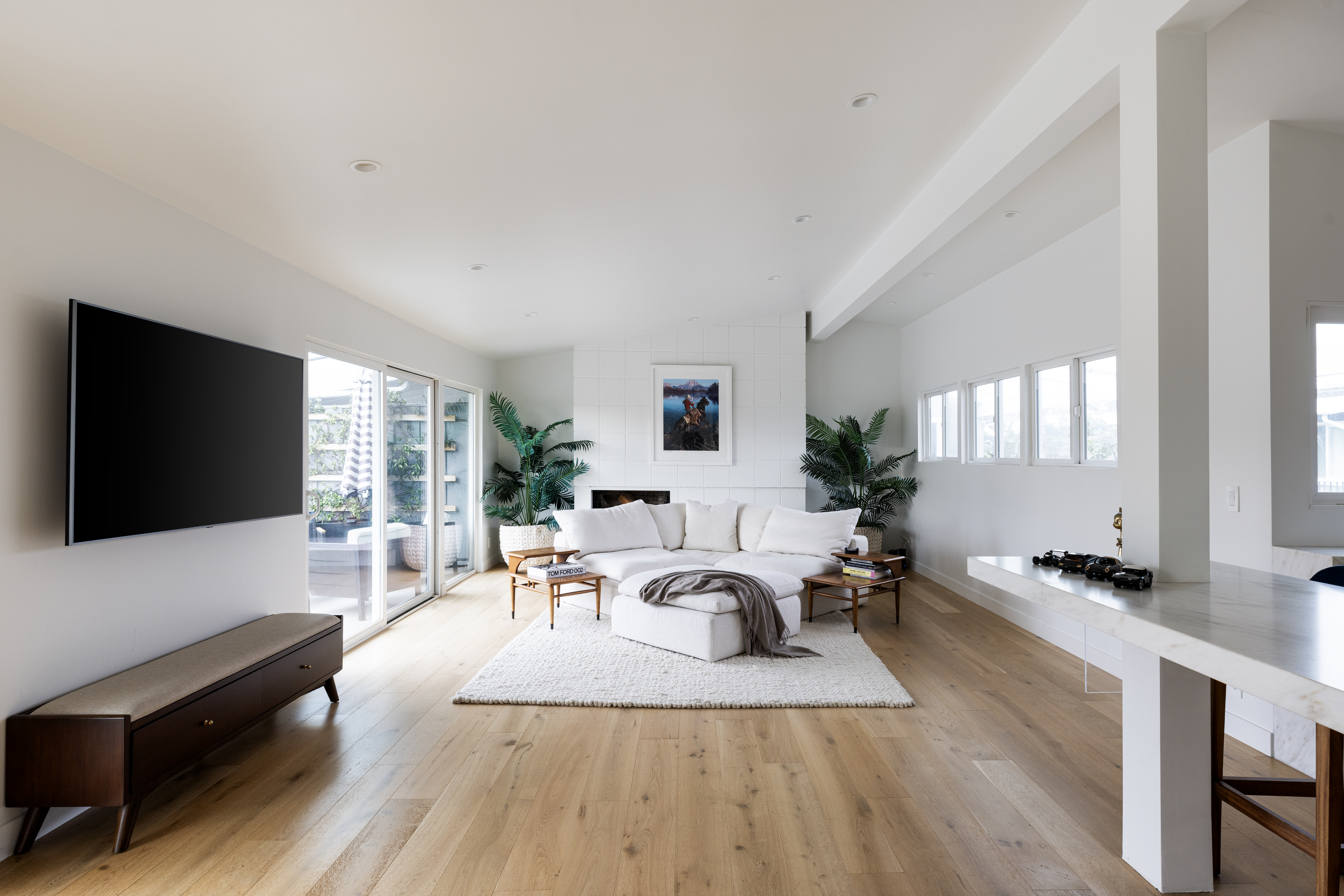 This bright and airy living room features light wood flooring and a neutral color palette, centered around a plush white sectional sofa and a matching ottoman. A tall, white-tiled fireplace surround serves as a focal point, flanked by large potted plants that add a touch of greenery. The space is illuminated by recessed lighting and natural light streaming through large glass sliding doors that open to an outdoor area.