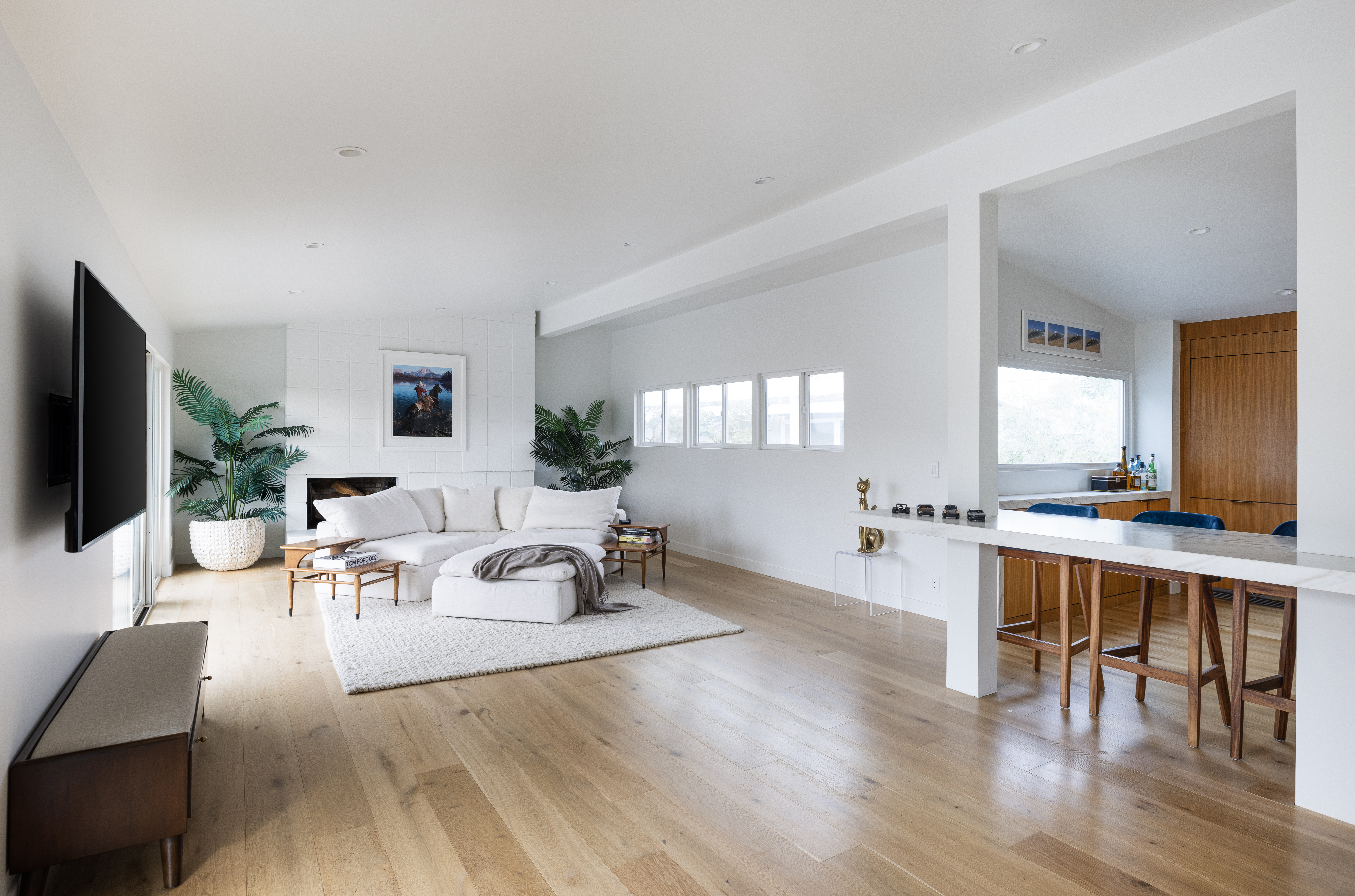 This bright, open-concept living area features light wood flooring and a neutral color palette that creates an airy, inviting atmosphere. A white sectional sofa sits atop a textured rug, facing a fireplace with a tall, white-tiled surround and a mounted television on the adjacent wall. To the right, a long, stone-topped counter serves as a breakfast bar, connecting the living space to the kitchen area, which is accented by warm wood cabinetry.