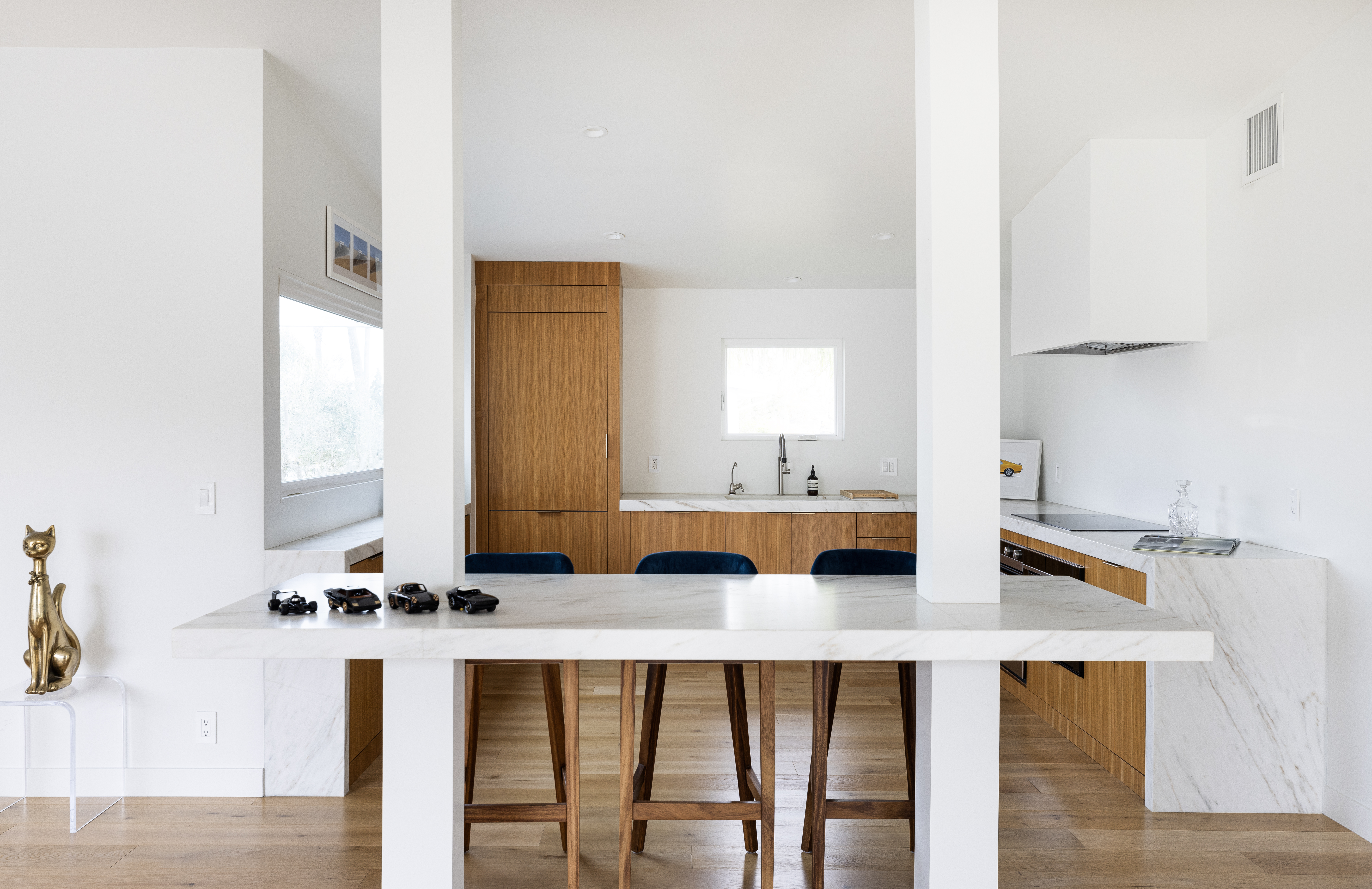This bright, modern kitchen features a central island with a stone countertop and three wooden bar stools, framed by two prominent white structural pillars. The space showcases a clean aesthetic with light wood cabinetry, integrated appliances, and a minimalist white range hood. A gold cat figurine sits on a clear pedestal in the foreground, adding a touch of personality to the sleek, open-concept design.