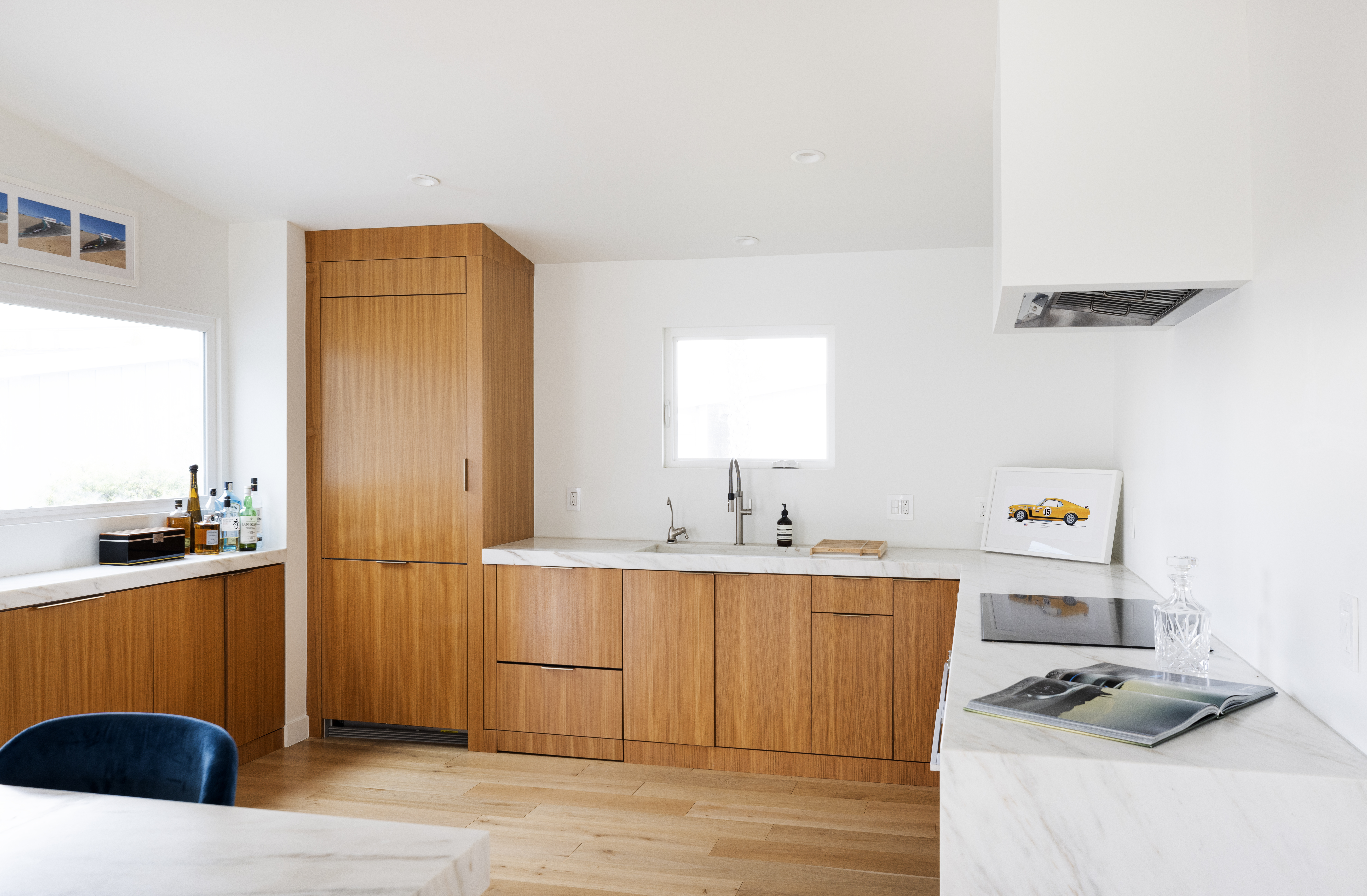 This modern kitchen features warm-toned wood cabinetry paired with light-colored stone countertops and a clean, minimalist aesthetic. The space includes a built-in refrigerator, a sleek cooktop, and a central sink area, all illuminated by recessed lighting and natural light from the windows. A framed car print adds a touch of personality to the bright, white-walled room, creating a sophisticated and inviting atmosphere.