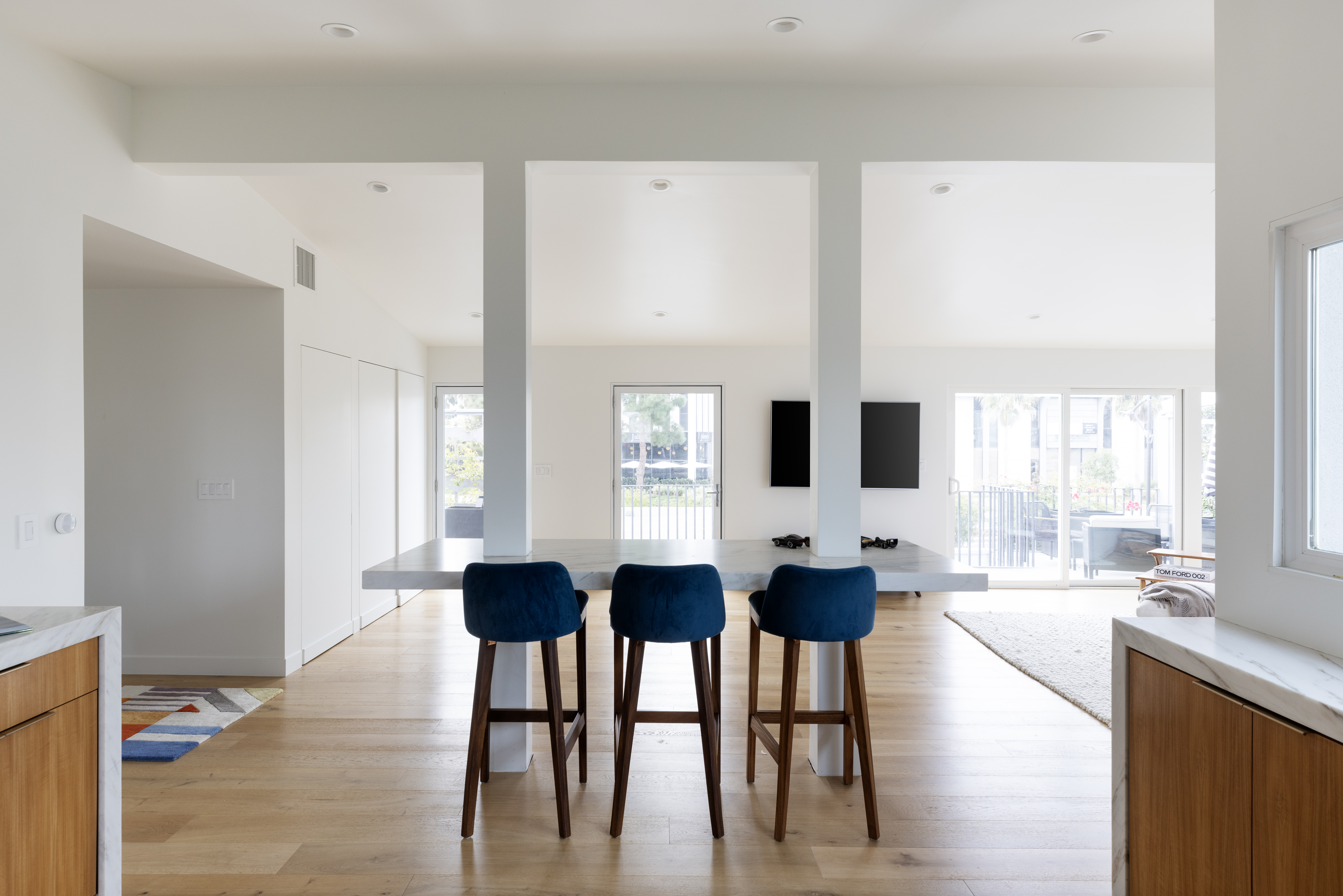 This bright, open-concept kitchen features a central island with a stone countertop and three blue upholstered bar stools. The space is defined by clean white walls, light wood flooring, and a minimalist aesthetic that flows into the adjacent living area. Large glass doors at the rear provide ample natural light and a view of the outdoor patio space.