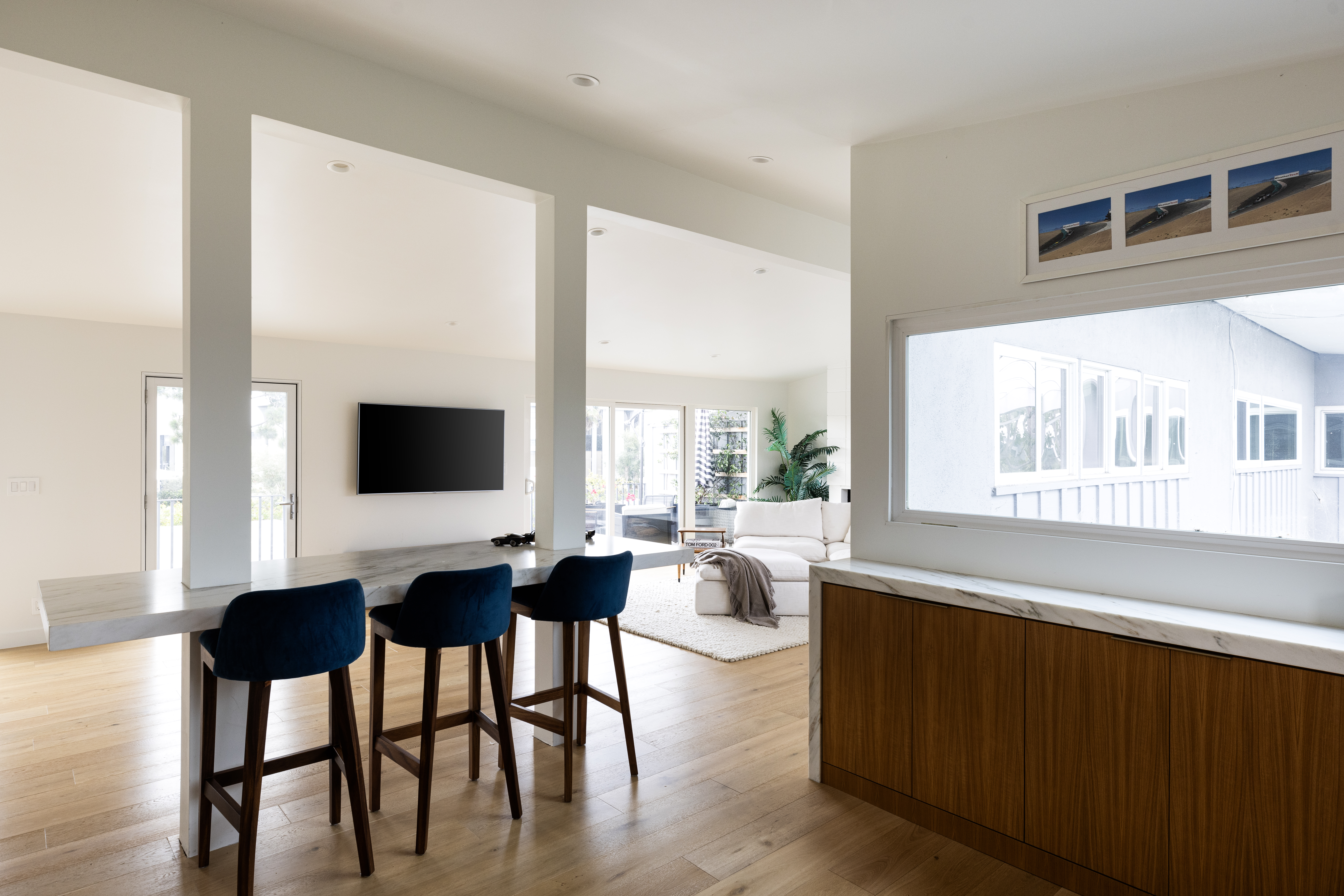 This open-concept interior features a sleek kitchen island with a stone countertop and three dark blue upholstered bar stools, serving as a transition point between the kitchen and the living area. The space is characterized by light wood flooring, white walls, and a minimalist aesthetic that flows into a bright living room with a white sofa. A large horizontal window on the right provides a view of the exterior, while the overall design emphasizes a clean, airy, and modern atmosphere.