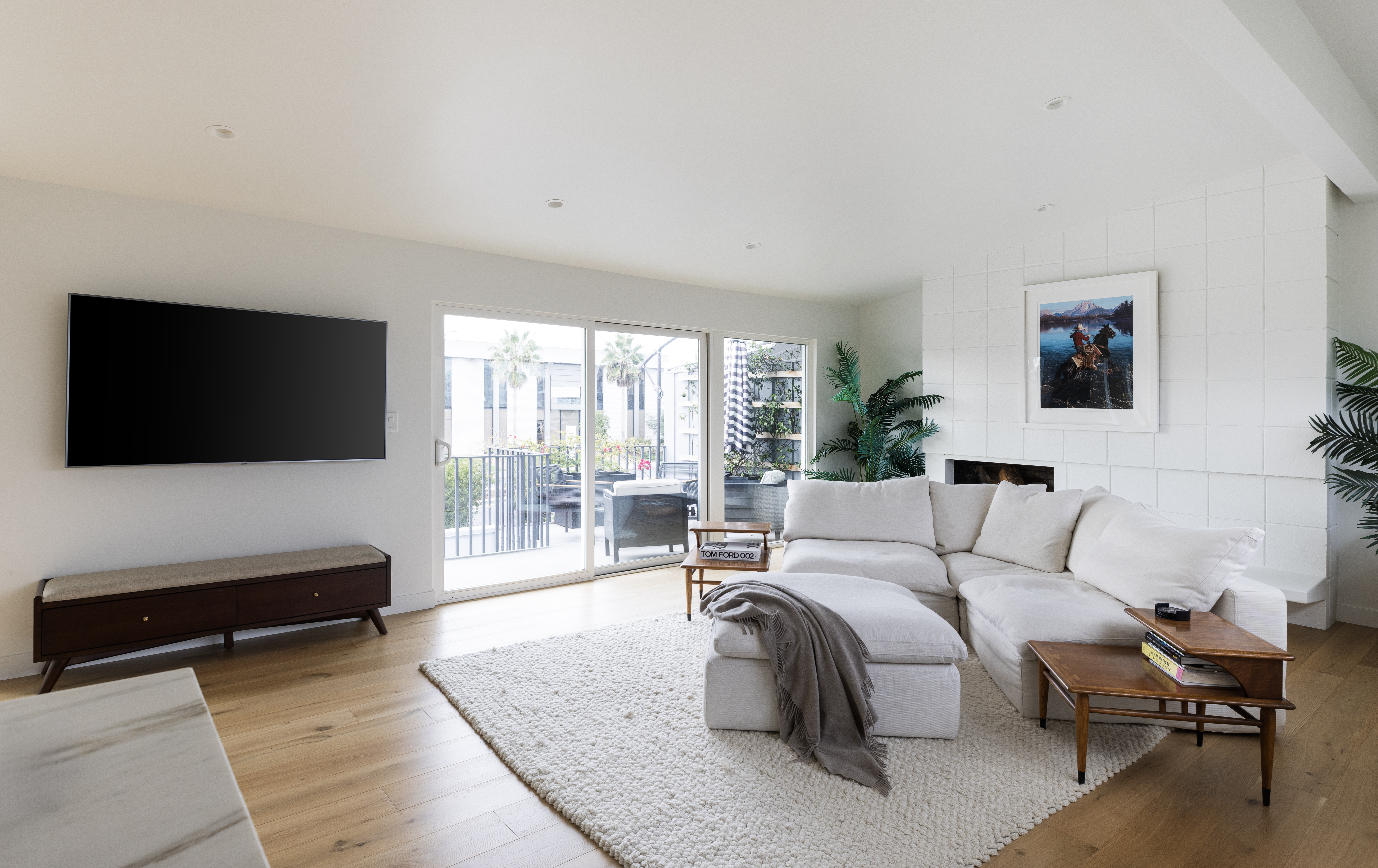 This bright and airy living room features a large white sectional sofa arranged around a textured area rug, creating a comfortable and inviting seating area. A dark wood media console sits beneath a wall-mounted television, while a fireplace with a white tiled surround serves as a focal point on the adjacent wall. Large sliding glass doors provide a seamless transition to an outdoor patio, filling the space with natural light and offering a view of the exterior.