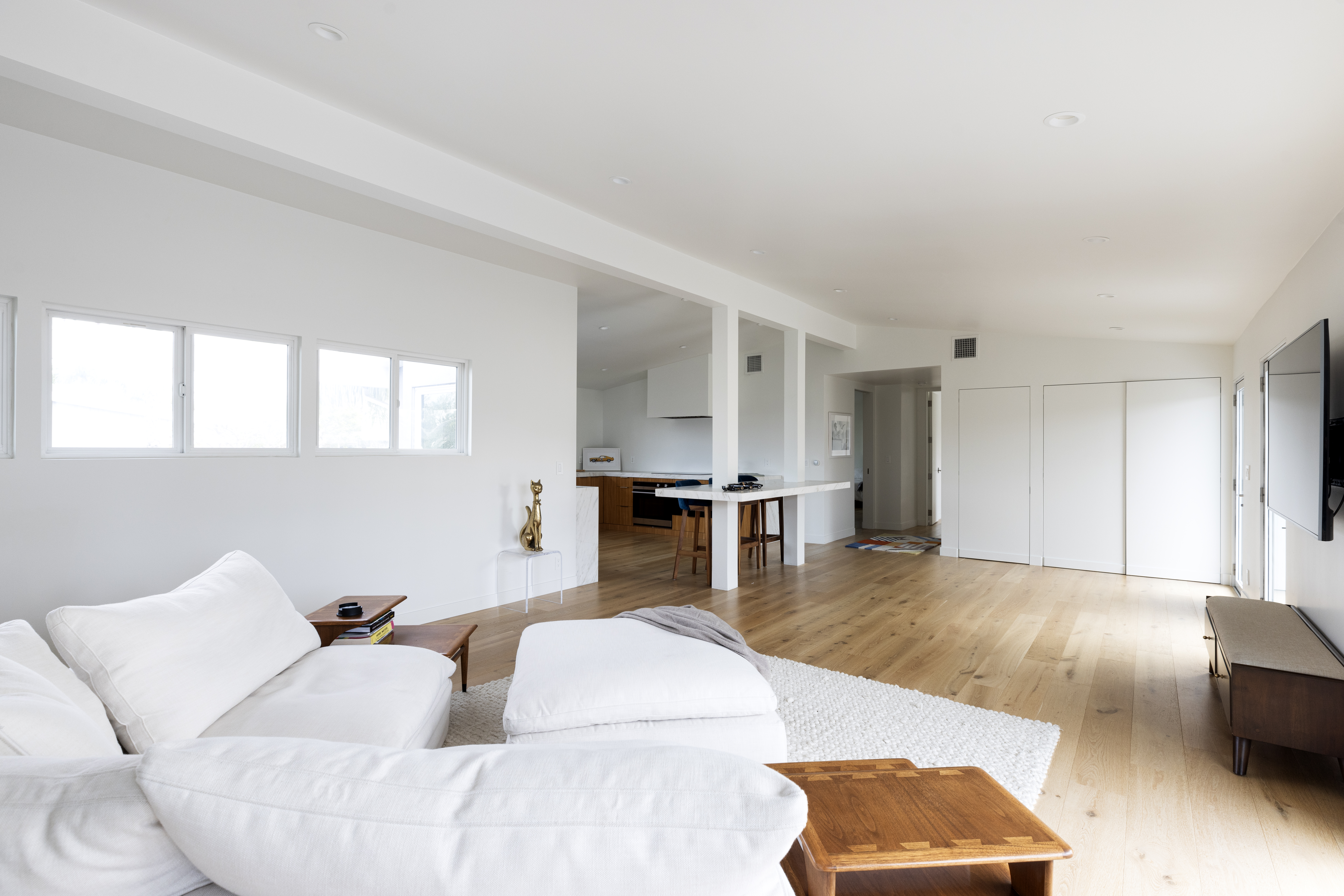 This bright and airy open-concept living space features light wood flooring and a neutral color palette, creating a clean, modern aesthetic. The room is anchored by a large white sectional sofa and a textured rug, with a view extending into a kitchen area featuring a central island and bar seating. The perspective is from the living area looking toward the kitchen, emphasizing the spacious, flowing layout of the home.