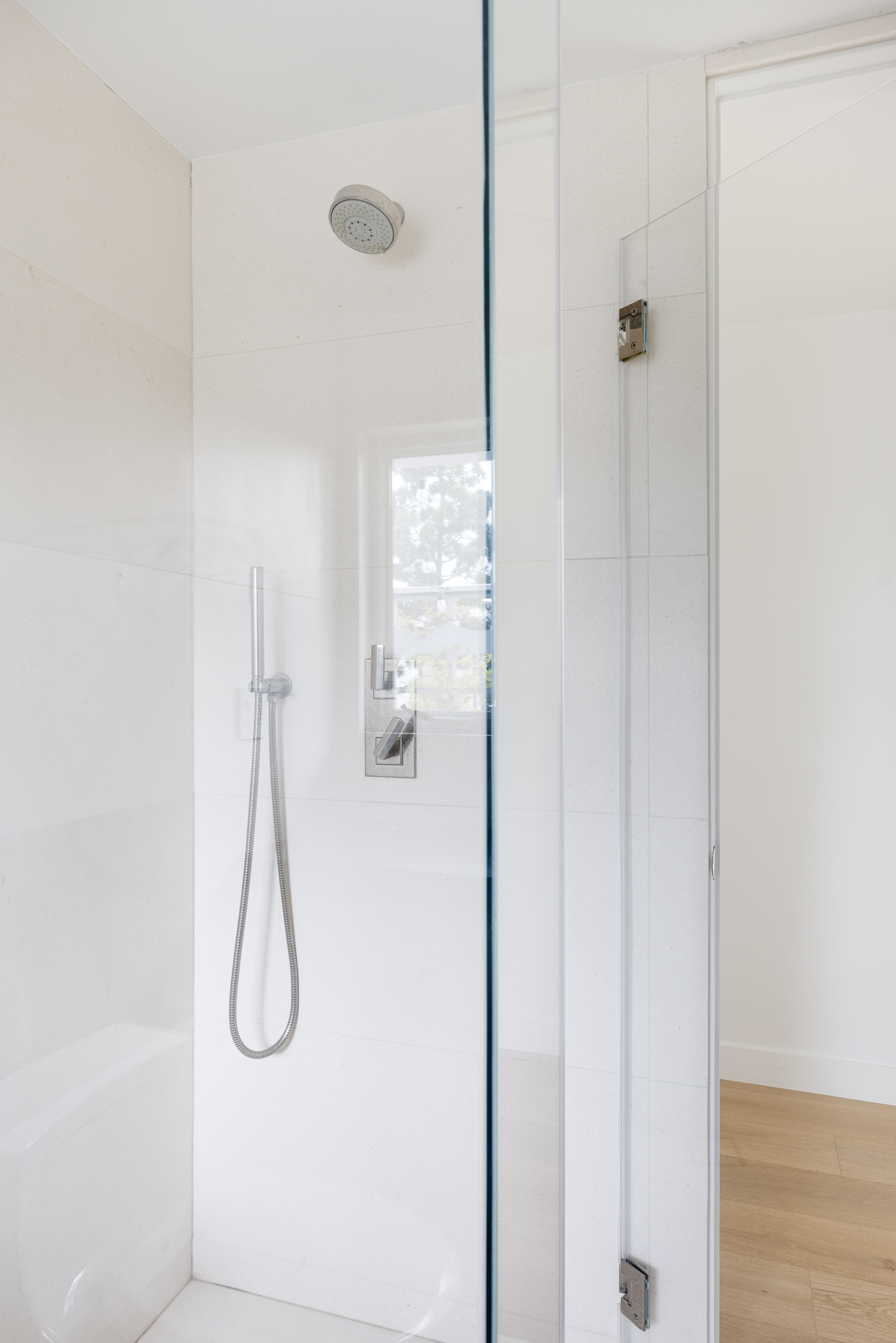 This image captures a modern, minimalist shower enclosure featuring a glass door and light-toned, large-format wall tiles. The shower is equipped with a wall-mounted rainfall showerhead and a separate handheld sprayer on a sliding rail, finished in polished chrome. The perspective looks directly into the shower stall, highlighting the clean lines and bright, airy aesthetic of the bathroom space.