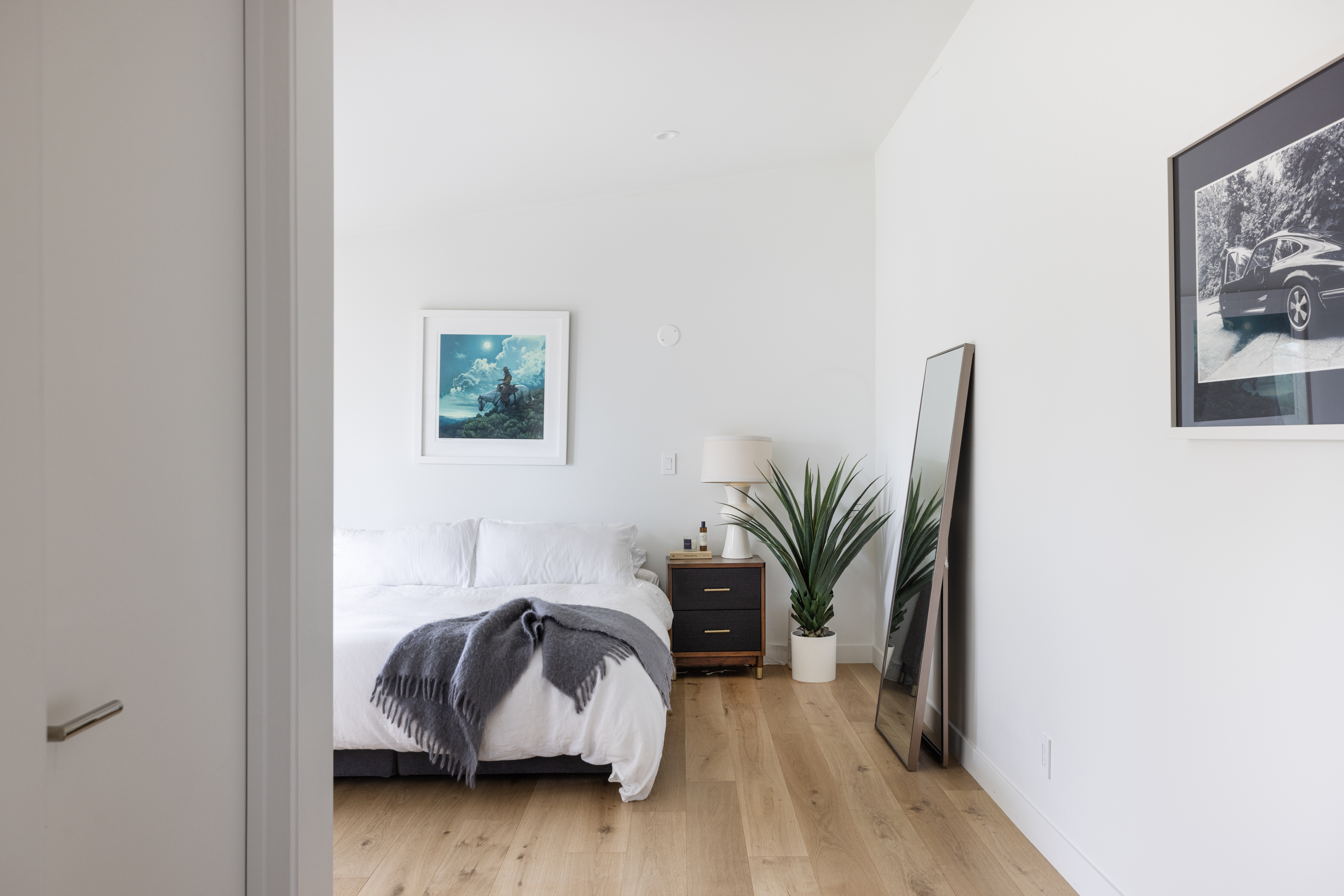 This bright and minimalist primary bedroom features light wood flooring and crisp white walls, creating an airy and serene atmosphere. The room is furnished with a bed dressed in white linens, a dark wood nightstand with a modern lamp, and a tall floor mirror leaning against the wall. A large potted plant adds a touch of greenery, while framed artwork on the walls provides a personalized, contemporary aesthetic.