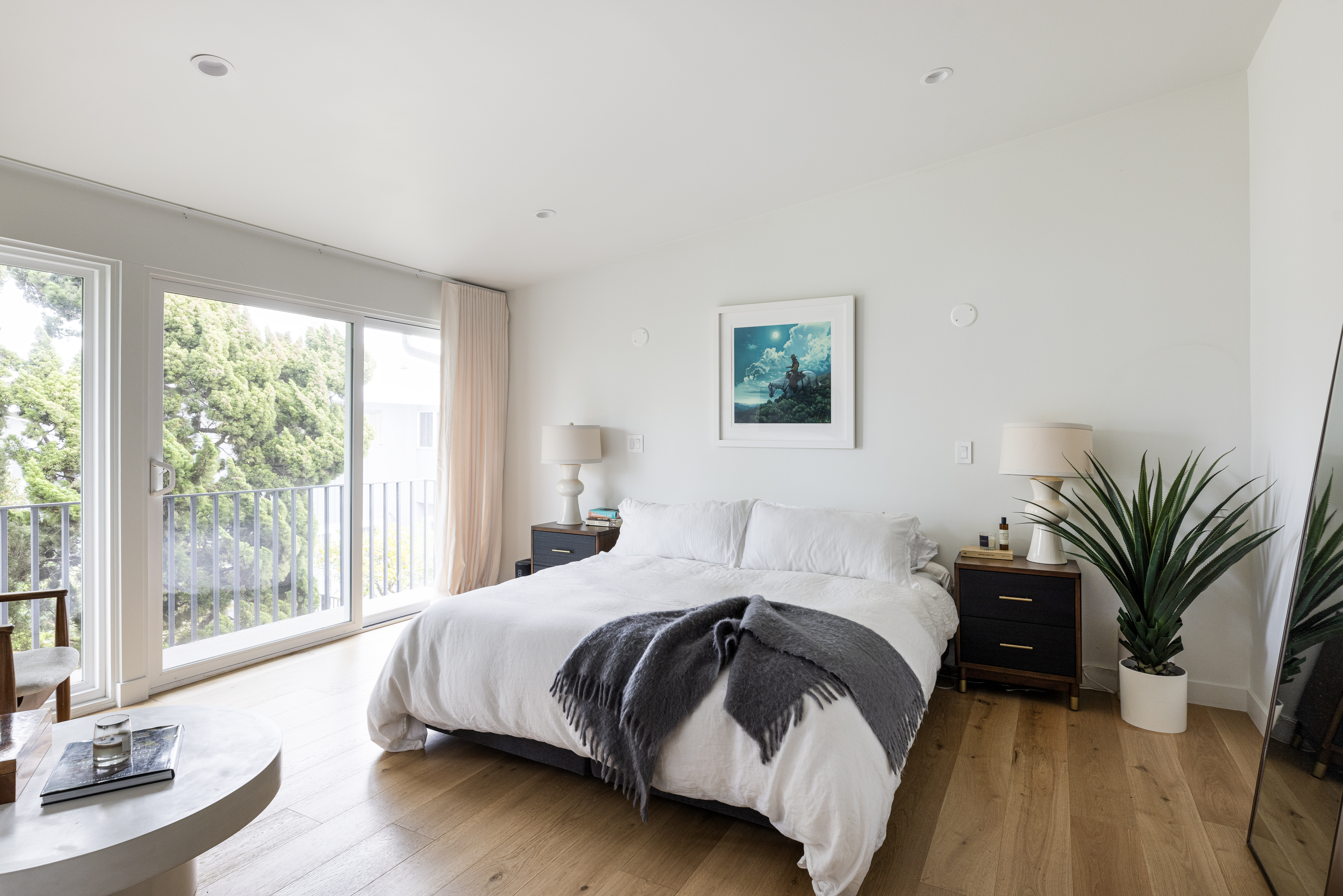 This bright and airy primary bedroom features light wood flooring and a neutral color palette, centered around a plush bed dressed in white linens with a dark grey throw blanket. A large sliding glass door provides access to a private balcony with a view of lush greenery, while dark wood nightstands and a potted plant add contrast to the space. The room is illuminated by recessed lighting and soft lamp light, creating a serene and inviting atmosphere.