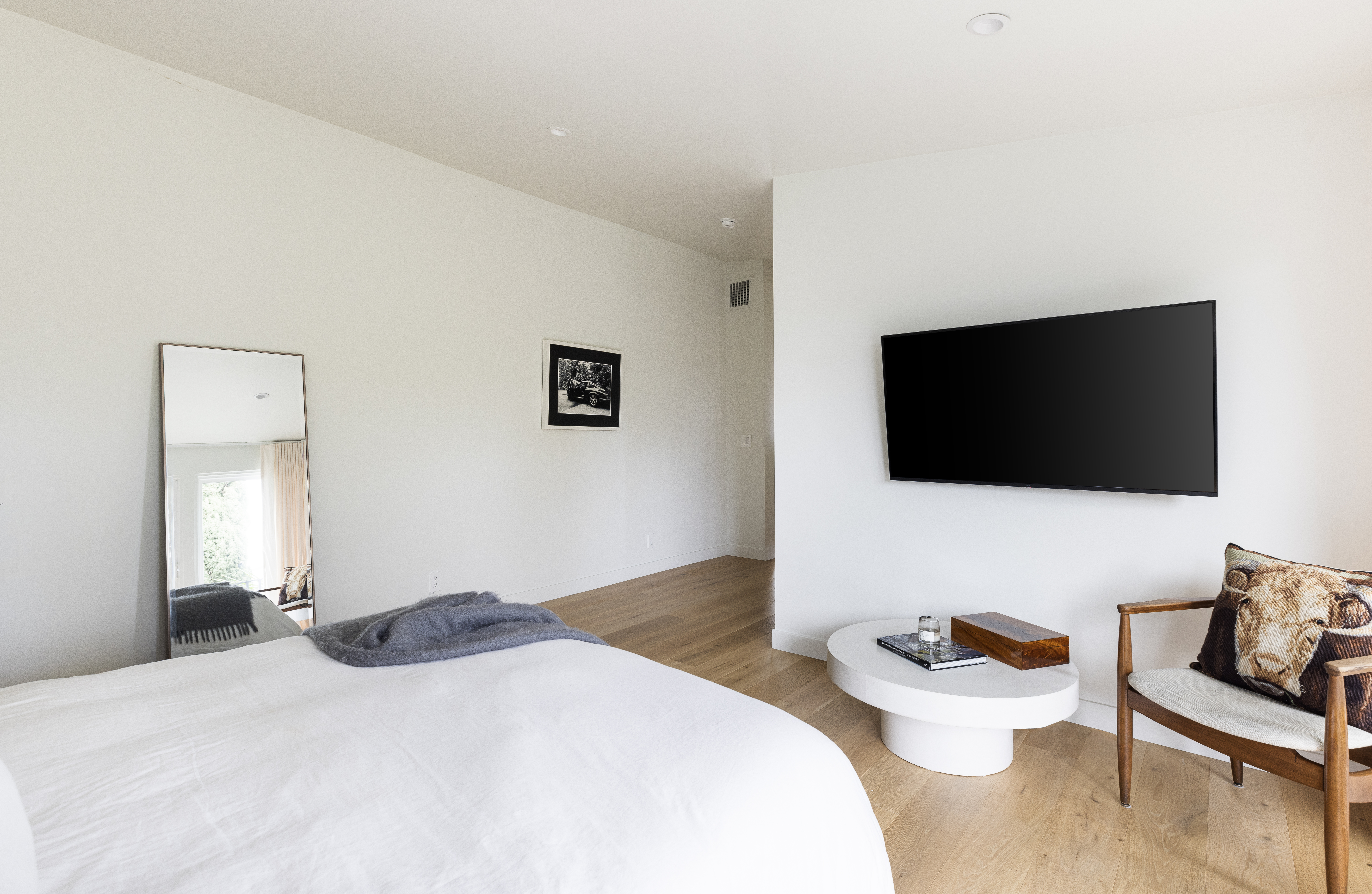 This bright and minimalist primary bedroom features light wood flooring and crisp white walls, creating an airy and serene atmosphere. The room is furnished with a bed topped with a grey throw, a modern white circular coffee table, and a wooden armchair accented with a patterned throw pillow. A large wall-mounted television and a full-length floor mirror add functional elements to the clean, contemporary design.