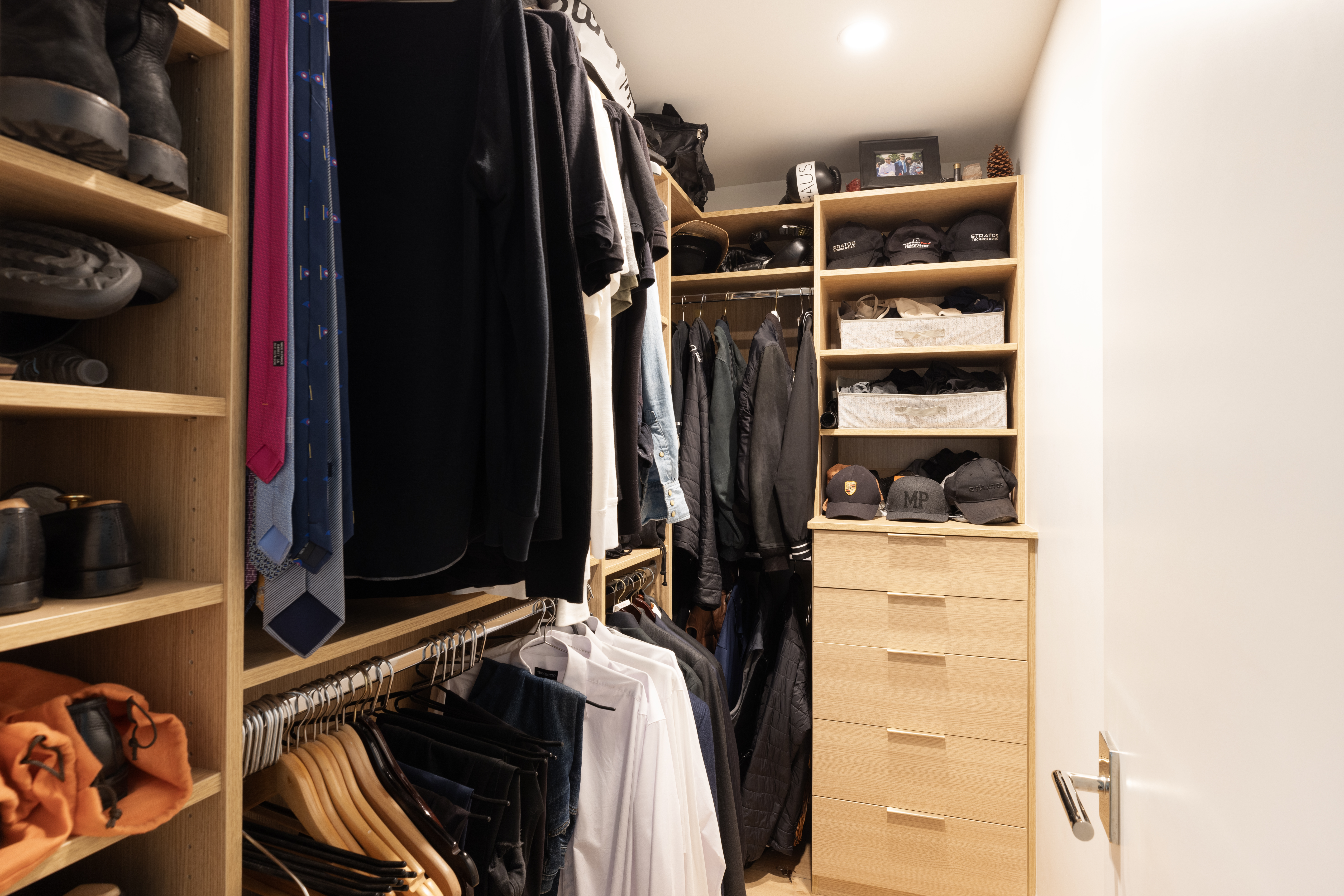 This organized walk-in closet features light wood shelving and cabinetry, providing ample storage for clothing, accessories, and hats. The space includes a hanging rod for shirts and jackets, a dedicated section for ties, and a built-in dresser with multiple drawers. The perspective is a straight-on view into the closet, highlighting the efficient use of space and clean, modern aesthetic.