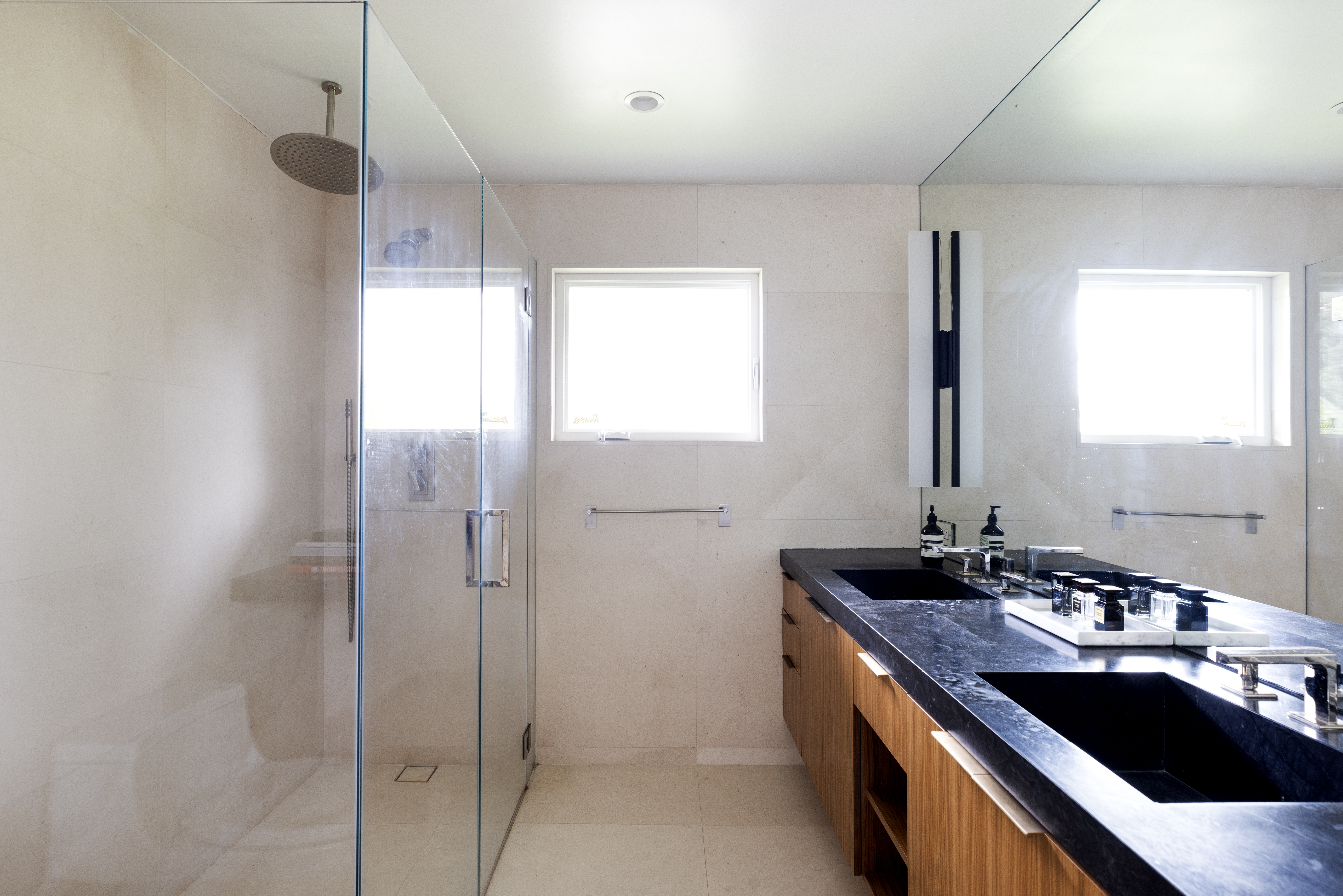 This modern bathroom features a sleek double vanity with a dark stone countertop and light wood cabinetry. A glass-enclosed walk-in shower with a rainfall showerhead sits to the left, while a large mirror reflects the space, enhancing the bright and airy atmosphere created by the window. The neutral color palette and clean lines provide a contemporary and sophisticated aesthetic.