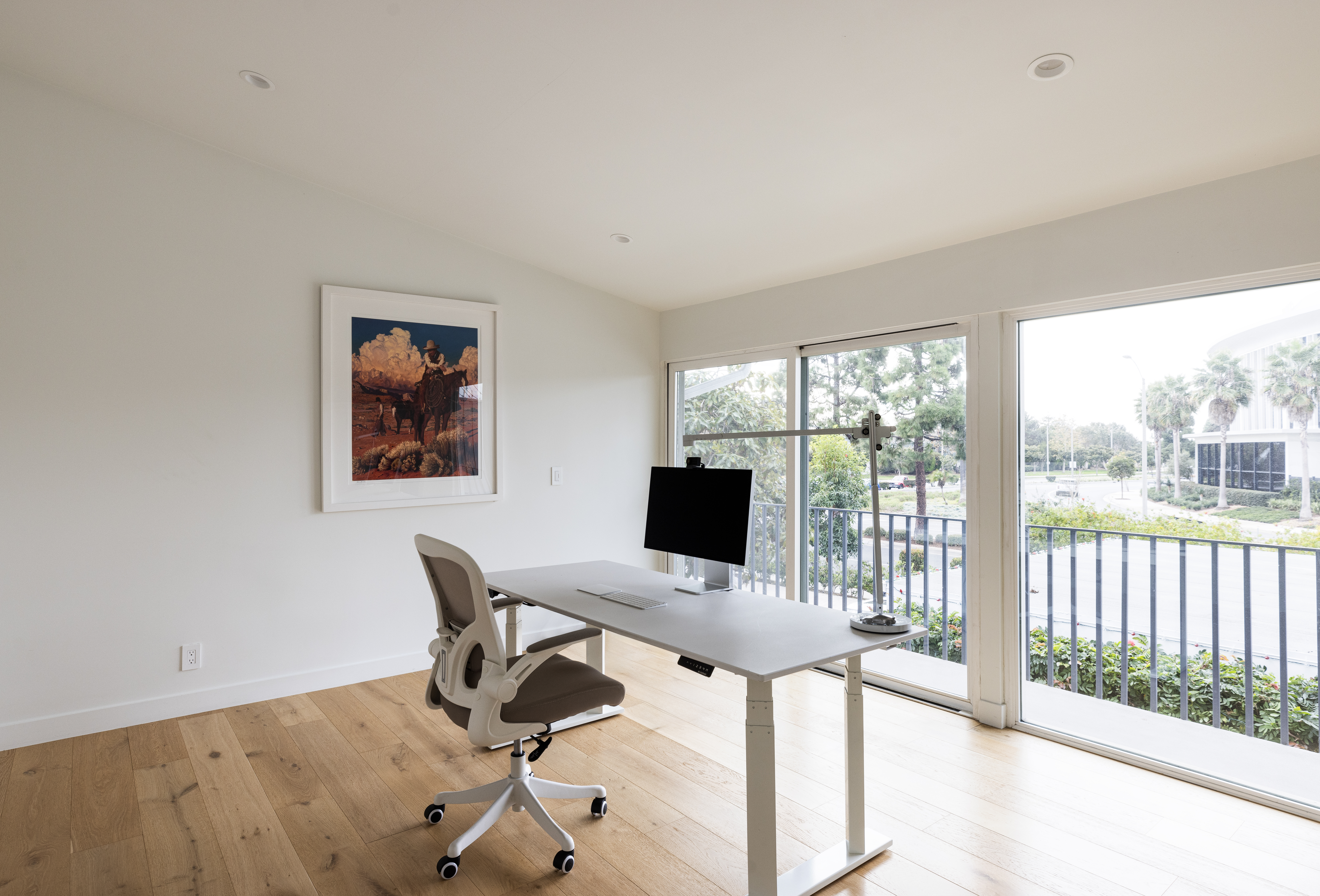This bright and minimalist home office features light wood flooring and a clean, white color palette. A modern adjustable-height desk is positioned to face large windows that provide ample natural light and a view of the outdoors, while a framed piece of art adds a touch of color to the wall. The space is uncluttered and professional, offering a serene environment for focused work.