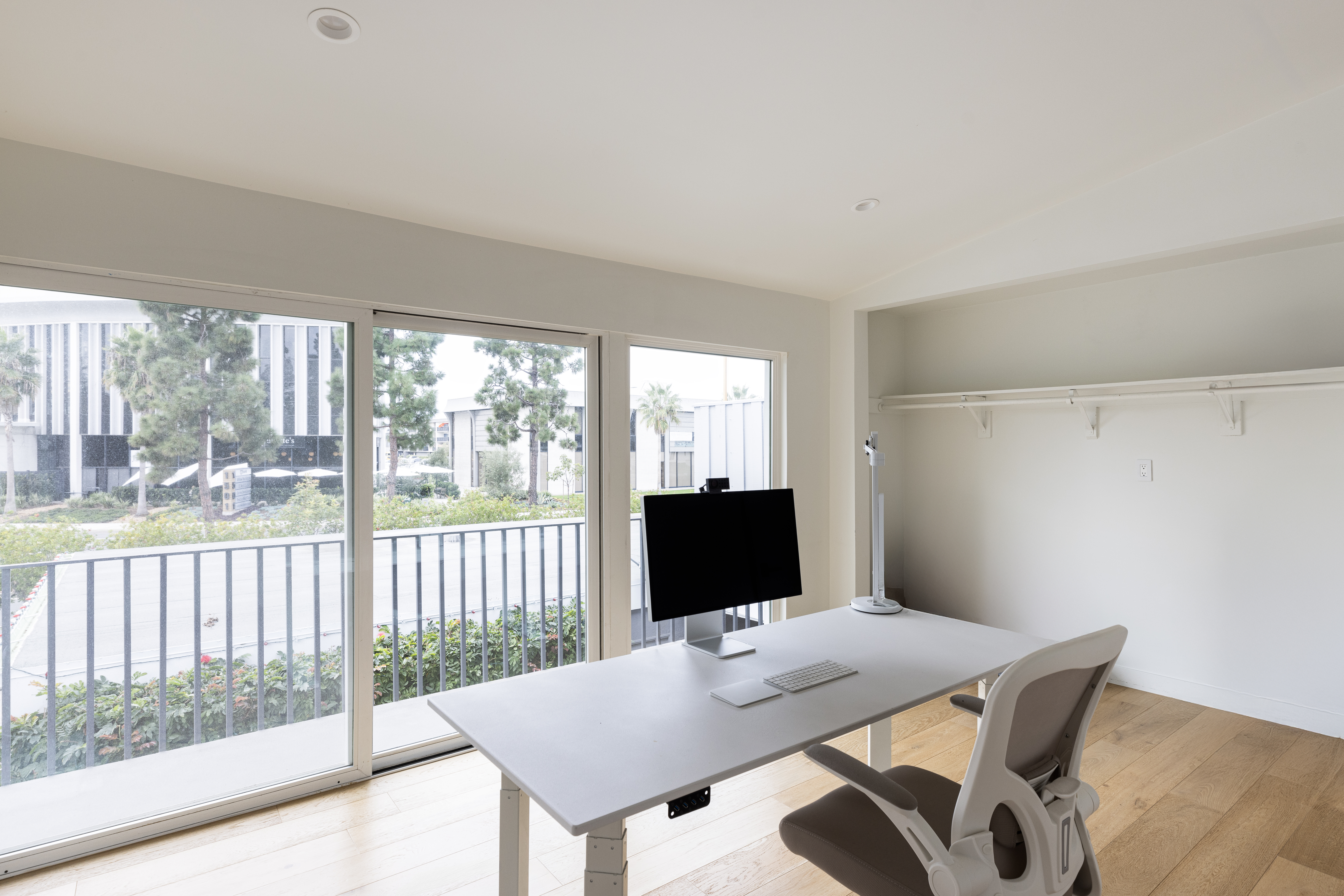 This bright and minimalist home office features a sleek, height-adjustable desk positioned in front of large sliding glass doors that offer an expansive view of the outdoors. The room is finished with light wood flooring and neutral walls, creating a clean and professional atmosphere. A modern ergonomic chair and a computer setup complete the workspace, while a recessed shelf area provides additional storage potential.
