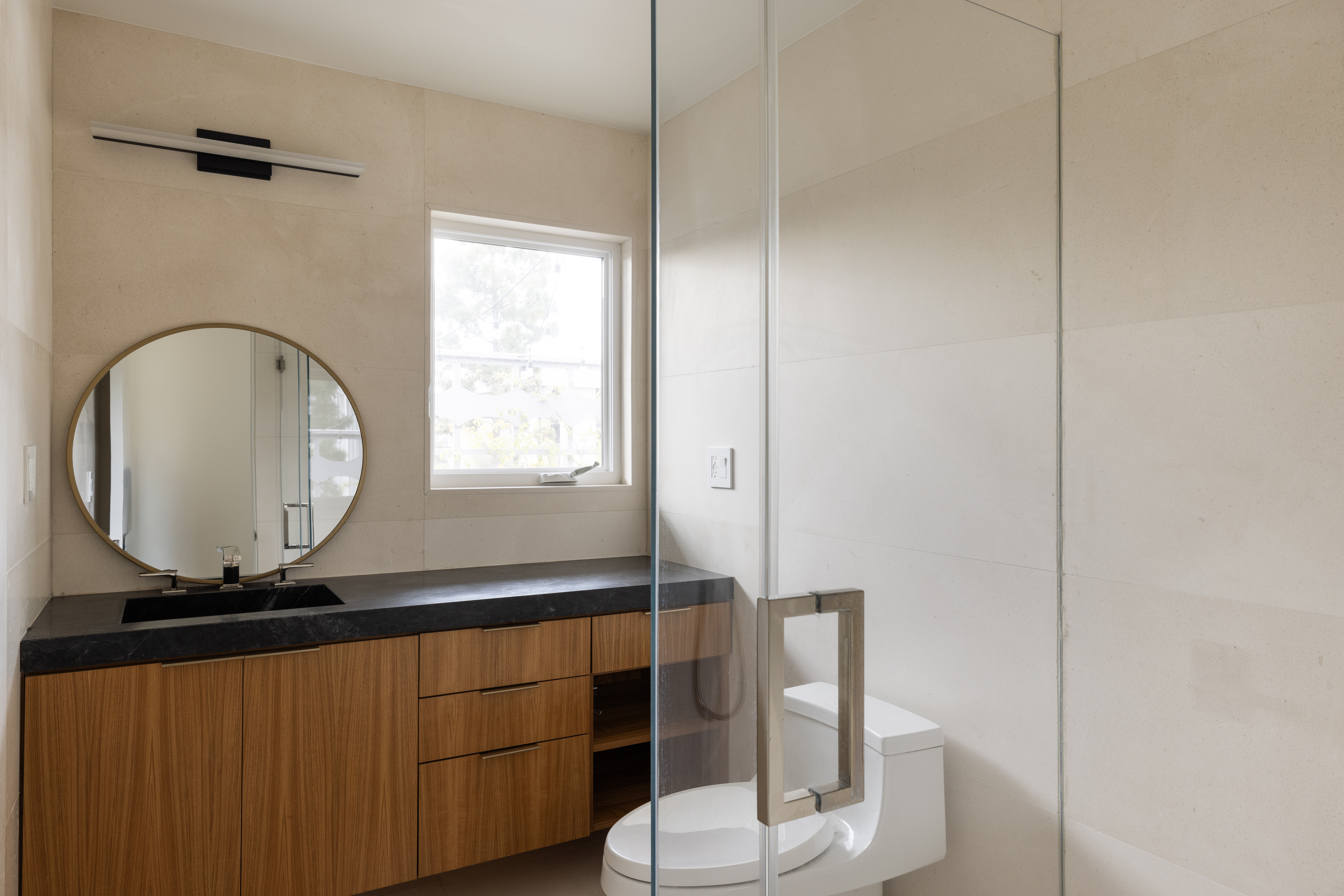 This modern bathroom features a sleek vanity with a dark stone countertop and light wood cabinetry, complemented by a circular gold-framed mirror. A glass-enclosed shower area sits adjacent to the vanity, and a window provides natural light to the space. The walls are finished with large, neutral-toned tiles, creating a clean and contemporary aesthetic.