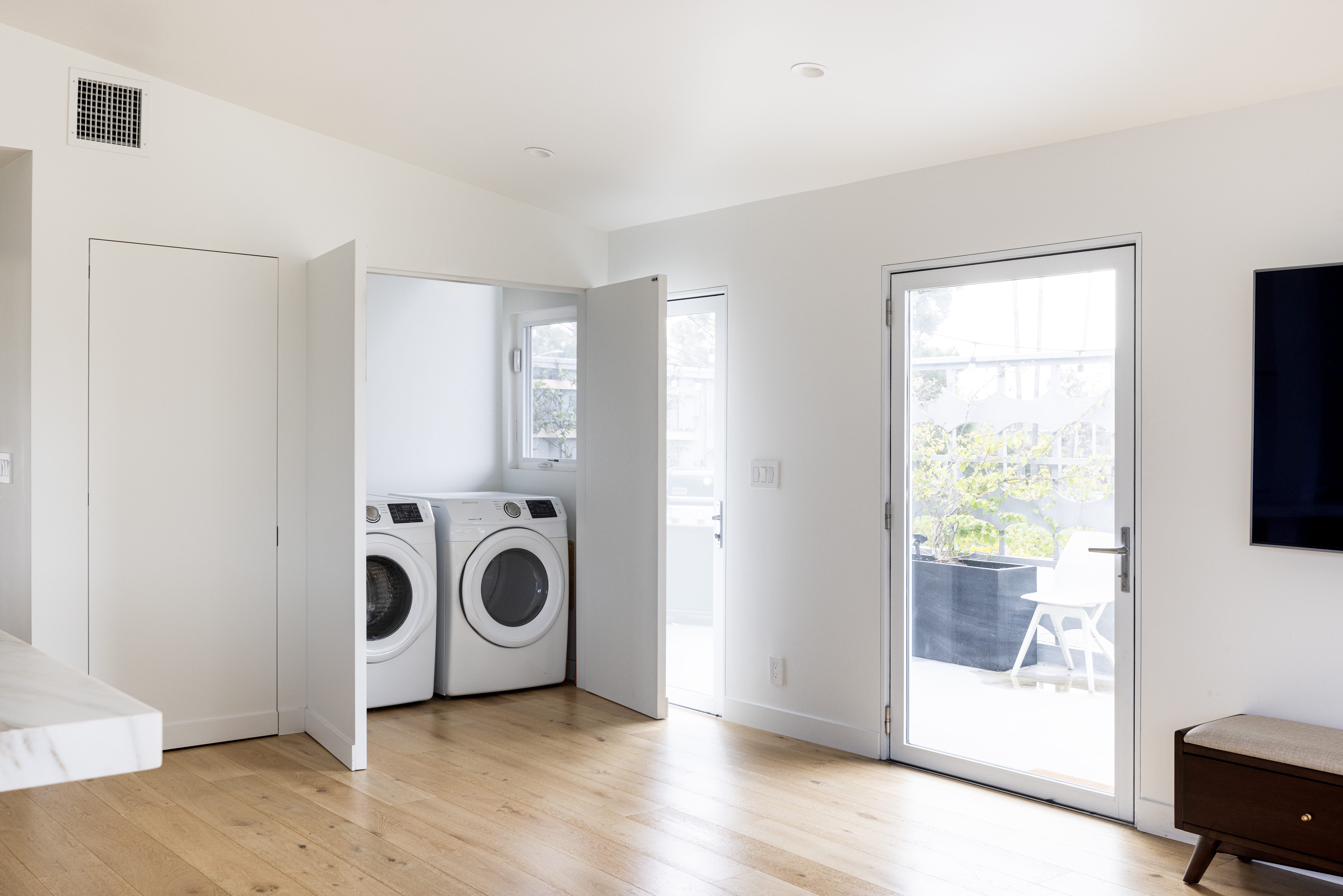 This bright, minimalist interior space features a laundry closet with a side-by-side washer and dryer set tucked behind open doors. The room is finished with light wood flooring and crisp white walls, leading to a glass-paned door that opens onto an outdoor patio area. A small dark wood bench sits to the right, while the clean, neutral aesthetic creates a functional and airy transition space.