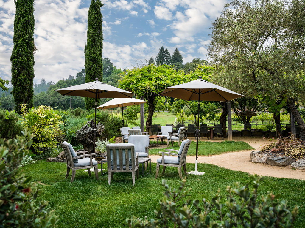 This serene garden setting features a lush green lawn furnished with comfortable outdoor seating arranged under large, neutral-toned umbrellas. A gravel pathway winds through the space, leading toward a shaded dining area nestled beneath a mature vine-covered arbor. The scene is framed by tall, slender cypress trees and expansive greenery, creating a peaceful and inviting atmosphere perfect for outdoor entertaining.