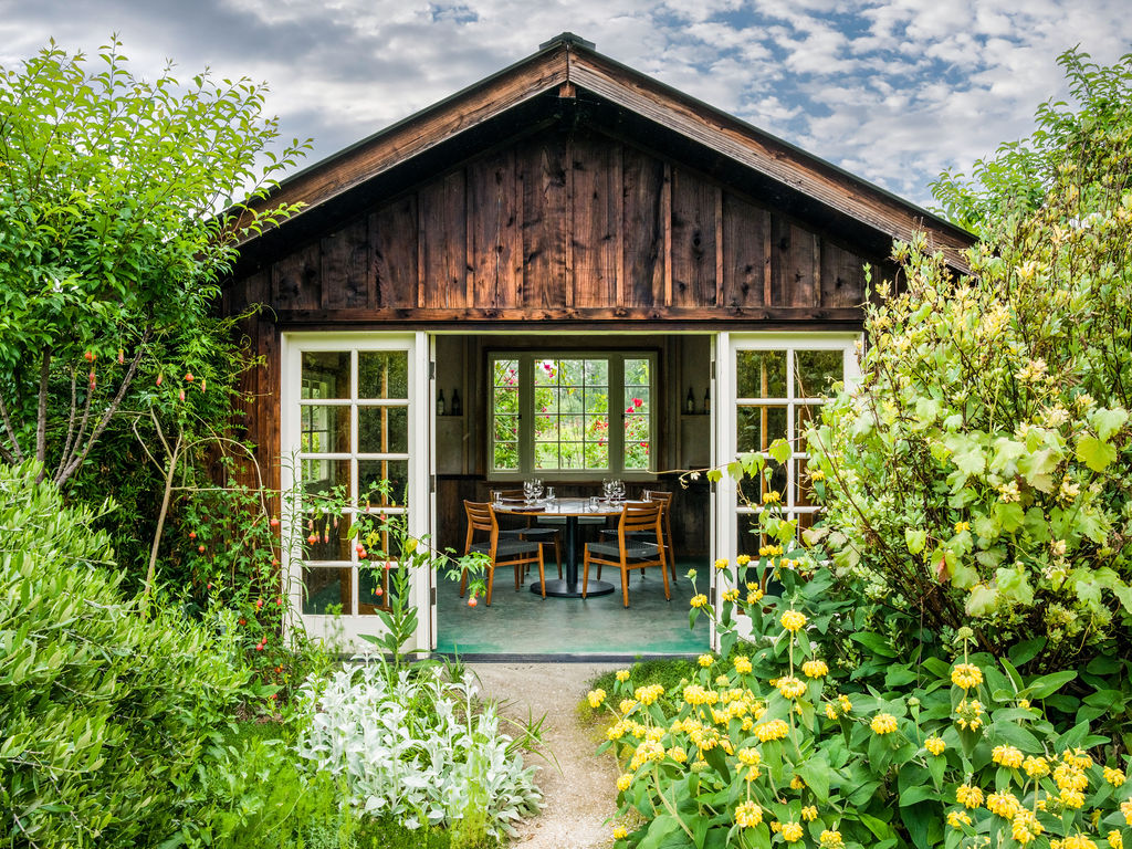 This charming, rustic garden structure features dark wood siding and wide-open French doors that reveal a cozy dining area inside. The scene is framed by lush, vibrant greenery and a foreground of bright yellow flowers, creating an inviting and serene atmosphere. The perspective is a straight-on, eye-level shot that highlights the seamless transition between the manicured garden and the intimate interior space.