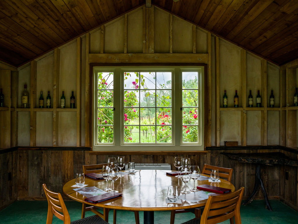 This rustic dining room features a circular wooden table set for a group, surrounded by wooden chairs and framed by wood-paneled walls. A large, multi-paned window serves as the focal point, offering a picturesque view of a lush garden filled with blooming roses. The space is accented with wine bottles displayed on built-in wall shelving, creating an intimate and charming atmosphere suitable for private tastings or gatherings.