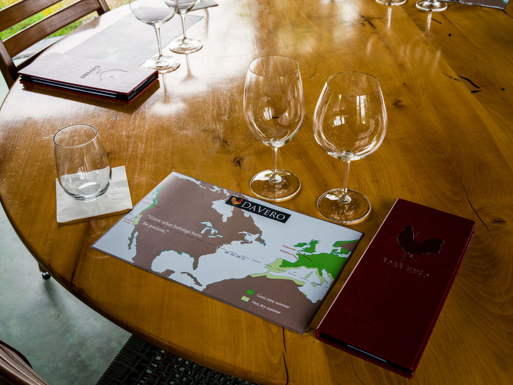 A high-angle, cinematic shot captures a polished wooden dining table set for a wine tasting experience. The table features several empty wine glasses, a small glass tumbler on a napkin, and two branded menus, one of which displays a map highlighting wine regions. The warm, natural wood grain of the table creates an inviting and sophisticated atmosphere suitable for a winery or upscale dining space.
