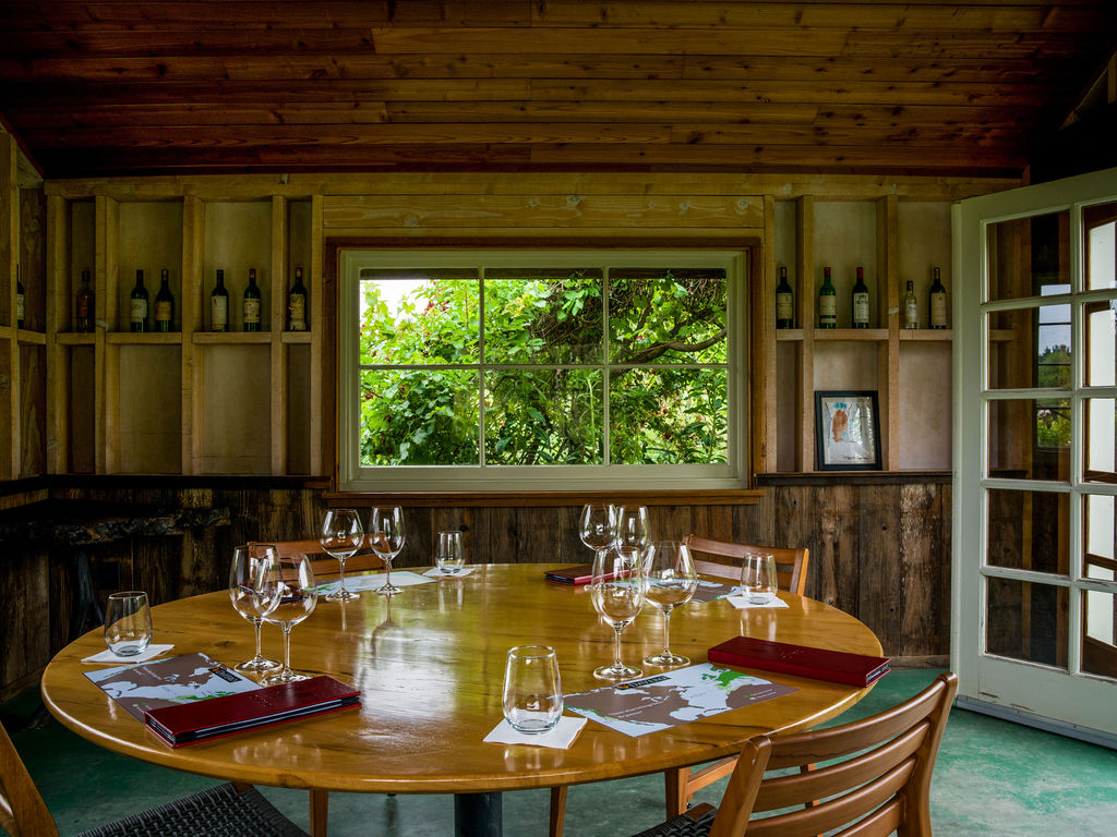 This rustic dining room features a large, circular light wood table set for a tasting experience, complete with wine glasses and menus. The walls are paneled in warm-toned wood, with built-in shelving displaying wine bottles on either side of a central window that looks out onto lush greenery. The space offers an intimate, inviting atmosphere, perfect for a private dining or wine-tasting setting.