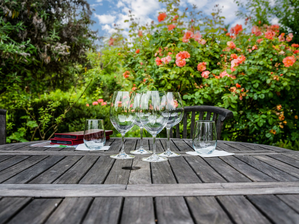 A rustic wooden circular table is set for an outdoor gathering, featuring several wine glasses and two water tumblers on white napkins. The table is positioned in a lush garden setting, surrounded by vibrant orange rose bushes and dense greenery under a partly cloudy sky. The perspective is at eye level, capturing a serene and inviting atmosphere perfect for al fresco dining.