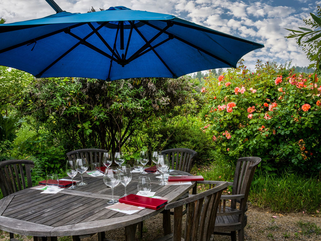 This outdoor dining area features a weathered wooden table set for a meal, positioned under a large, vibrant blue umbrella. The setting is nestled within a lush, blooming garden filled with vibrant orange roses and dense greenery, creating a serene and private atmosphere. The perspective is at eye level, capturing the inviting arrangement of glassware and red menus against the backdrop of a bright, cloudy sky.