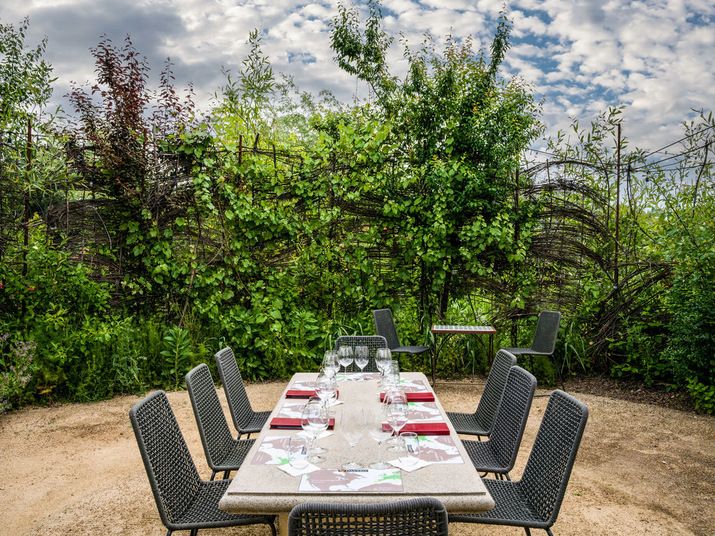 This outdoor dining area features a long, stone-topped table surrounded by dark woven chairs, set against a backdrop of lush greenery and a rustic woven-branch fence. The setting is framed by a gravel patio, creating an inviting and serene space for al fresco meals. The perspective is eye-level, capturing the intimate arrangement of the table settings and the natural, garden-like atmosphere.
