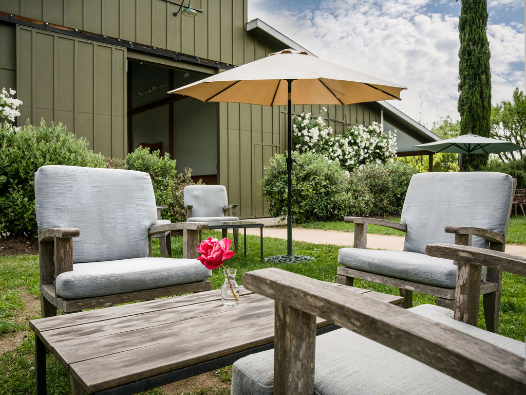This outdoor seating area features a rustic wooden coffee table and matching armchairs with light-colored cushions arranged on a lush green lawn. A large beige patio umbrella provides shade, while a vibrant pink flower in a glass vase adds a pop of color to the center table. In the background, a green barn-style structure with sliding doors and blooming white bushes creates a serene, pastoral setting.