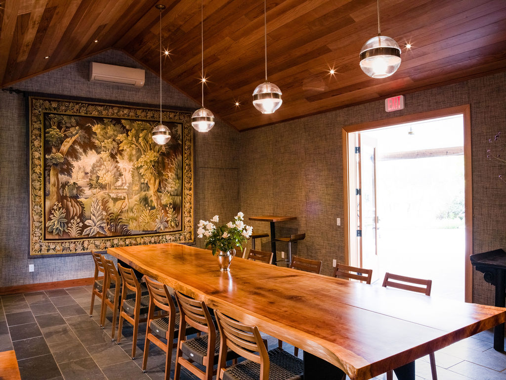 This dining room features a long, natural-edge wood table as the centerpiece, surrounded by wooden chairs. The space is accented by a large, ornate tapestry on the wall and a warm wood-paneled ceiling with four modern globe pendant lights. A doorway leads to an outdoor area, and the room is finished with textured wall coverings and dark stone flooring.