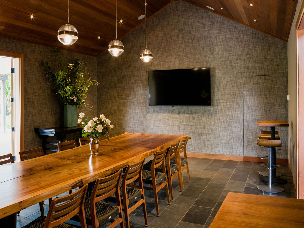 This sophisticated dining room features a long, live-edge wood table as the centerpiece, surrounded by multiple wooden chairs with woven seats. The space is defined by a vaulted wood-paneled ceiling, textured wall coverings, and slate-style flooring, creating a warm and rustic atmosphere. Three modern glass pendant lights hang above the table, while a wall-mounted television and a small bar area with stools complete the functional layout.