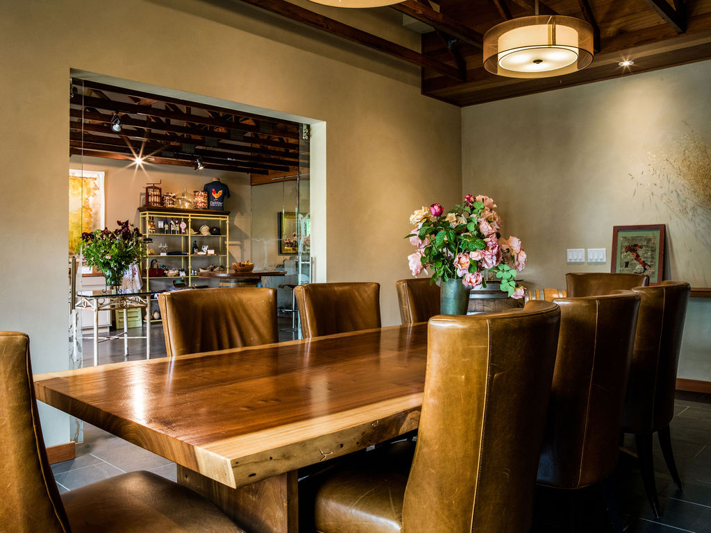 This elegant dining room features a substantial live-edge wood table surrounded by high-backed leather chairs, creating a warm and inviting atmosphere. A large floral arrangement serves as a centerpiece, while a view through a wide opening reveals an adjacent retail or display space with shelving. The room is illuminated by a modern drum pendant light, and the neutral walls provide a sophisticated backdrop for the rich textures of the furniture.