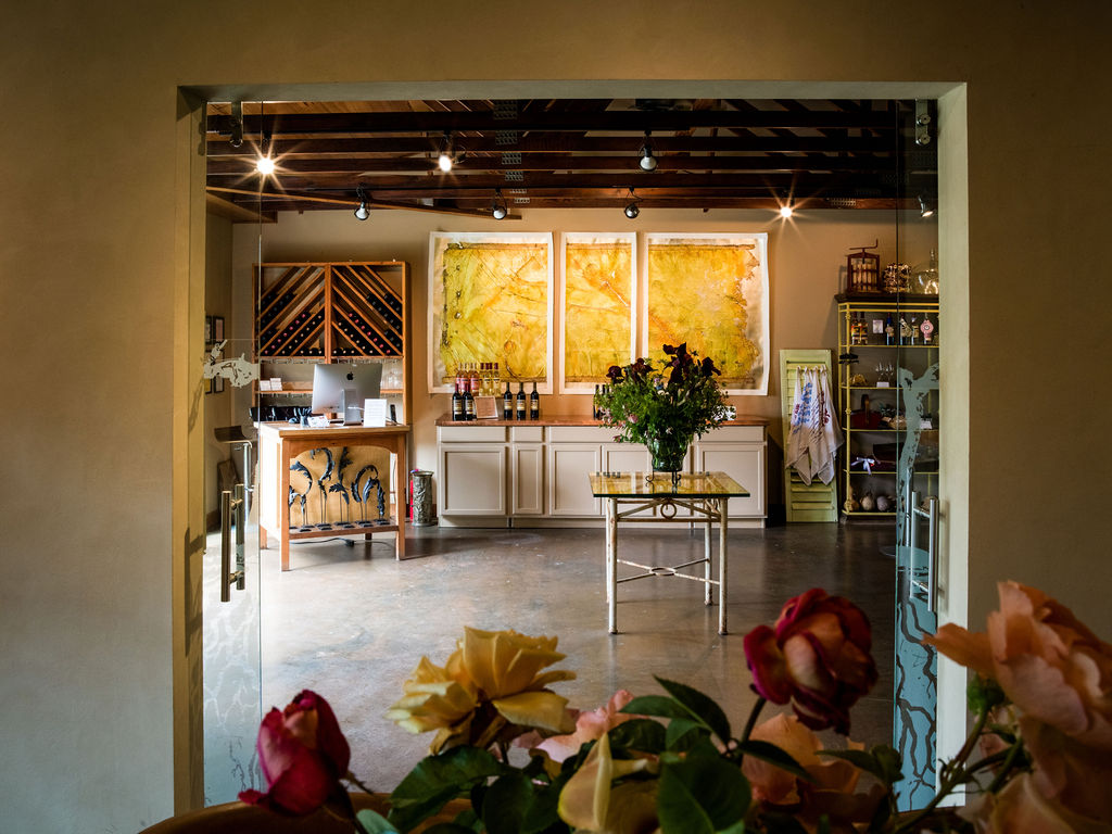 This inviting interior space, likely a wine tasting room or boutique retail area, is viewed through a glass doorway framed by soft-focus roses in the foreground. The room features a central glass-topped display table, a service counter with a computer, and three large, warm-toned abstract artworks mounted on the back wall. Exposed wooden ceiling beams and track lighting add a rustic yet professional aesthetic to the space.