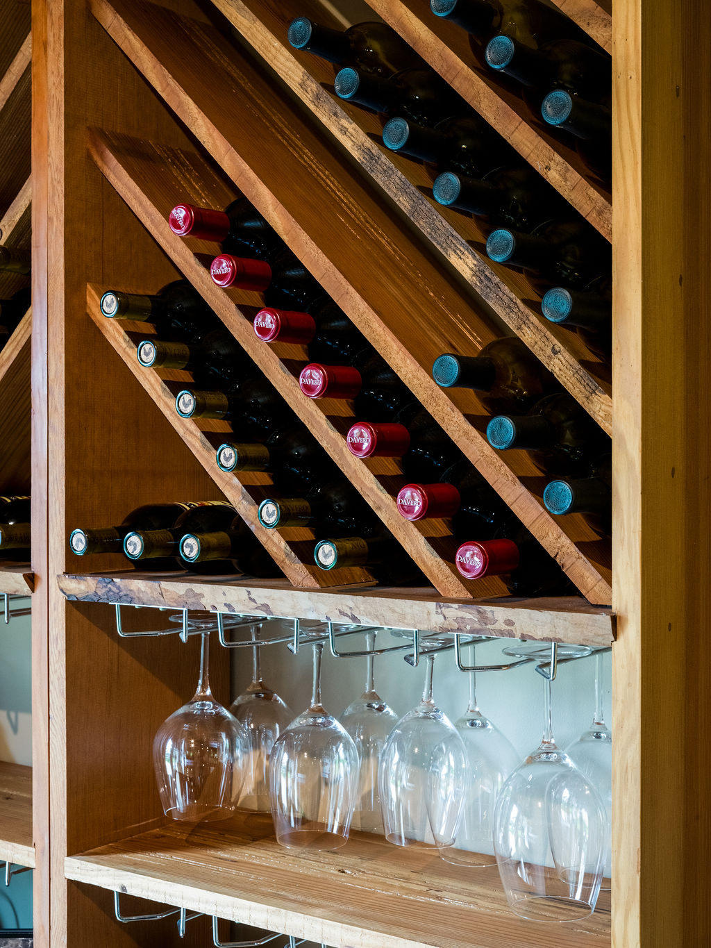 This close-up view highlights a custom-built wooden wine rack integrated into a home bar or cellar area. The rack features diagonal shelving for bottle storage, with several bottles visible, and a built-in rack below for hanging wine glasses. The warm-toned wood construction provides a rustic and sophisticated aesthetic suitable for a dedicated beverage storage space.