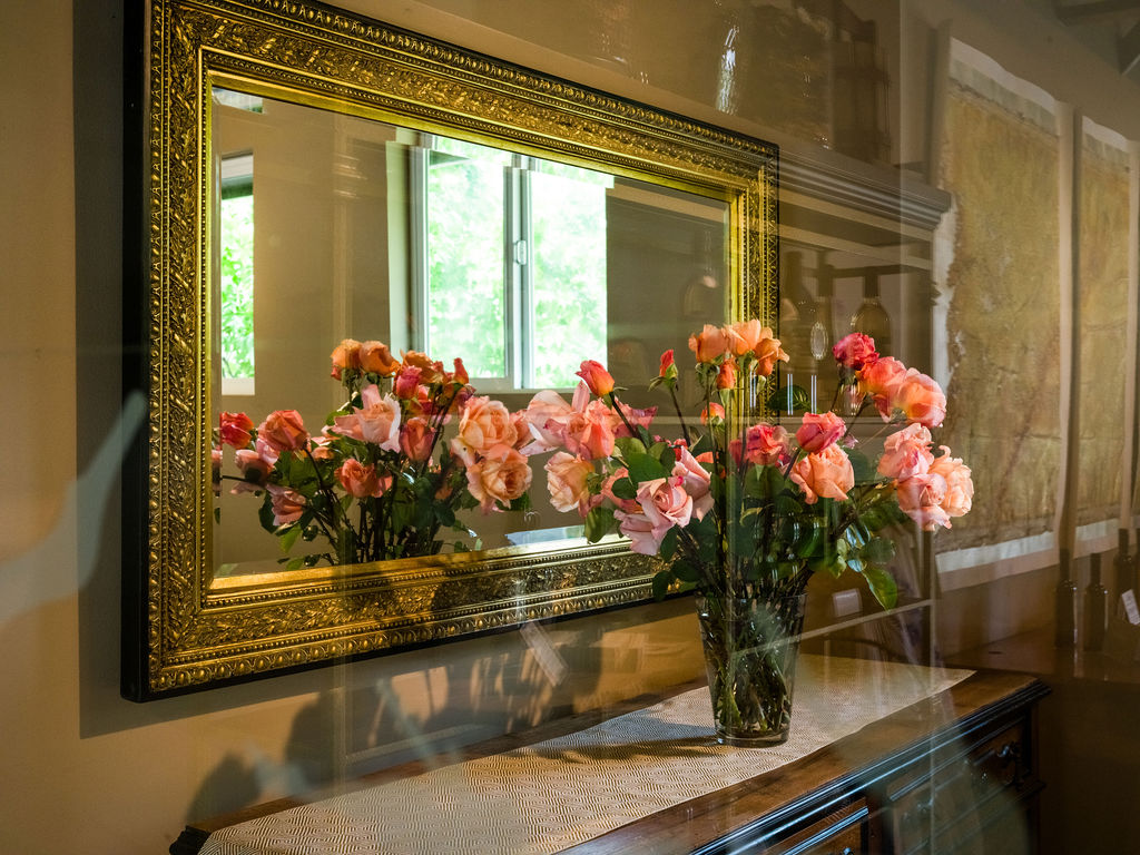 This interior detail shot features a large, ornate gold-framed mirror mounted above a wooden console table. A glass vase filled with fresh pink and peach roses sits on the table, creating a soft, elegant focal point that is reflected in the mirror. The composition captures a warm, inviting atmosphere, highlighting the decorative elements and the reflective quality of the space.