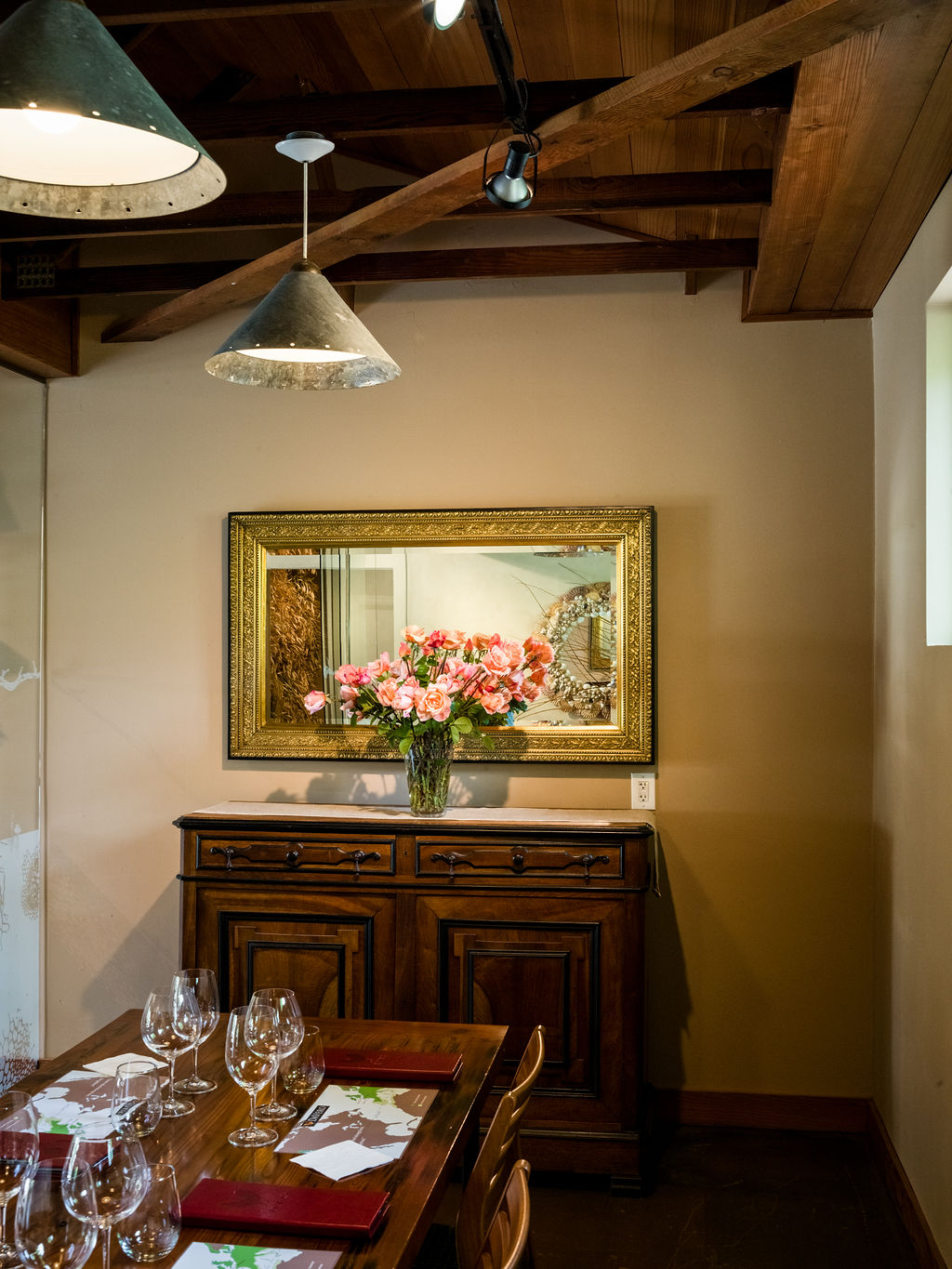 This dining room features a rustic aesthetic with exposed wooden ceiling beams and industrial-style pendant lighting. A dark wood sideboard sits against a neutral wall, topped with a large, ornate gold-framed mirror and a vibrant bouquet of pink roses. The foreground shows a portion of a wooden dining table set with wine glasses and menus, creating an inviting atmosphere for a meal.