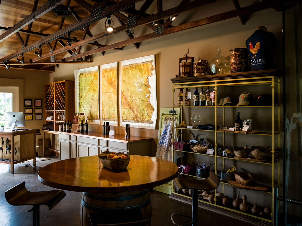 This interior space functions as a rustic tasting room or retail area, featuring a prominent round wooden table set atop a wine barrel base. The room is characterized by exposed wooden ceiling beams, warm ambient lighting, and a large yellow metal shelving unit displaying various merchandise and wine bottles. Three large, textured art pieces hang on the wall above a long service counter, creating a warm and inviting atmosphere typical of a winery or boutique farm shop.