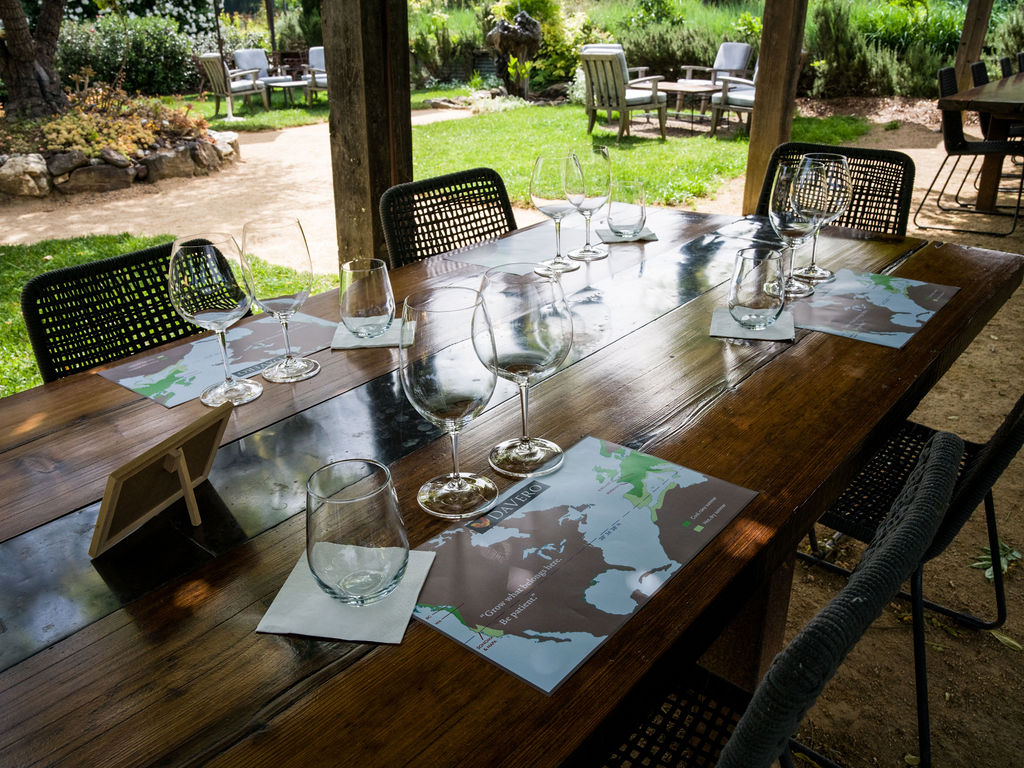This inviting outdoor dining area features a long, rustic wooden table set for a wine tasting or meal, complete with wine glasses and placemats. The space is sheltered by a wooden structure, providing a shaded view of a lush, landscaped garden with mature trees and additional seating areas. The setting offers a serene, open-air atmosphere perfect for entertaining or relaxing in a vineyard-style environment.