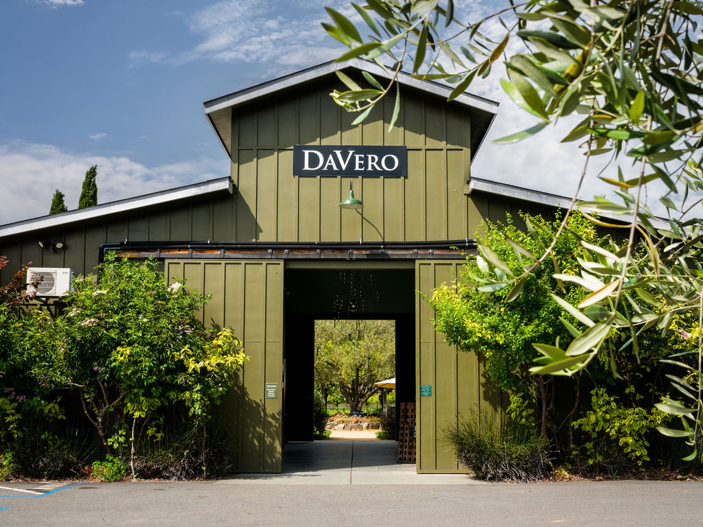 This image captures the front entrance of a rustic, olive-green barn-style building identified as DaVero. The structure features vertical siding, a prominent central gable, and a wide, open doorway that provides a framed view through to the lush, sunlit landscape beyond. The scene is framed by natural greenery and olive tree branches in the foreground, creating an inviting and serene atmosphere.