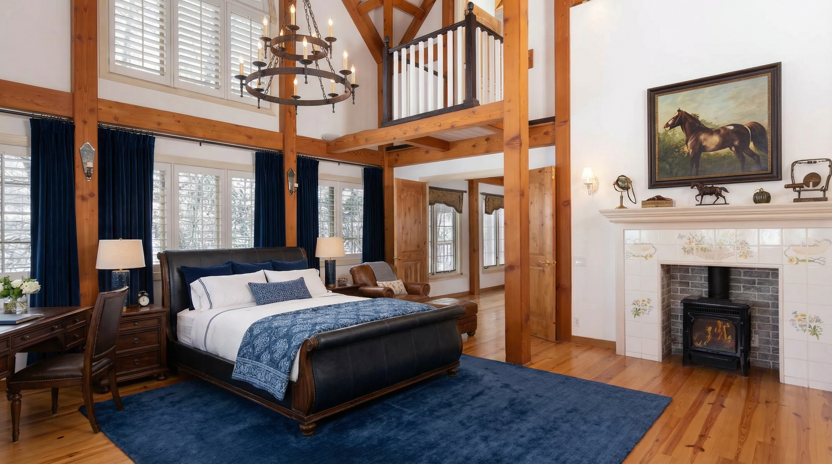 This spacious primary bedroom features dramatic vaulted ceilings with exposed timber beams and a loft overlook. The room is anchored by a dark leather sleigh bed and a decorative fireplace with painted tile work, set against warm wood flooring and deep blue accents. Large windows with plantation shutters fill the space with light, creating a rustic yet elegant retreat.