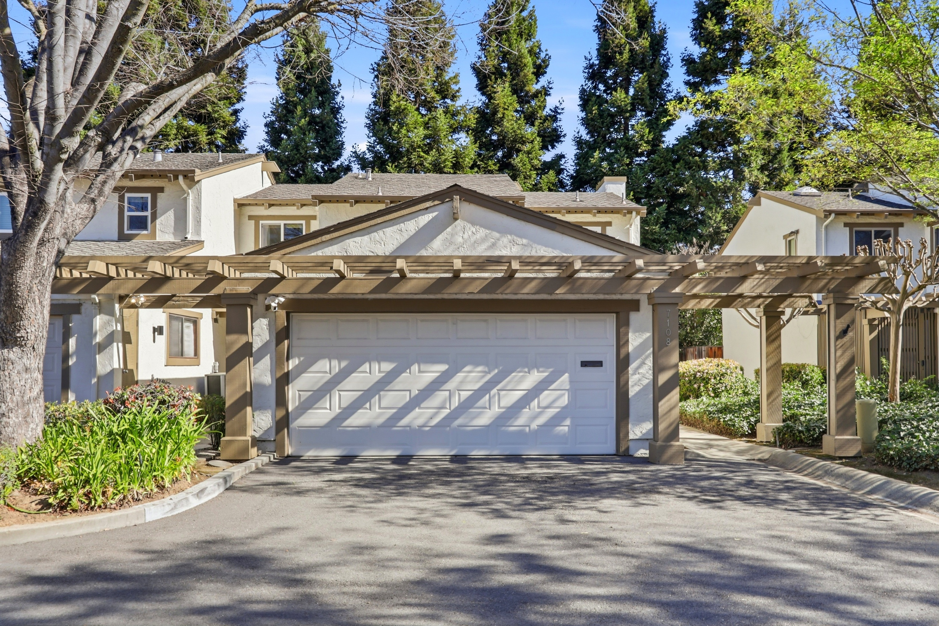 This image captures the front exterior of a residential property, focusing on a prominent two-car garage sheltered by an extensive wooden pergola structure. The home features a light-colored stucco facade with contrasting brown trim, set against a backdrop of mature evergreen trees and a clear blue sky. The perspective is a straight-on, eye-level shot that emphasizes the architectural symmetry and the inviting, well-maintained curb appeal of the residence.