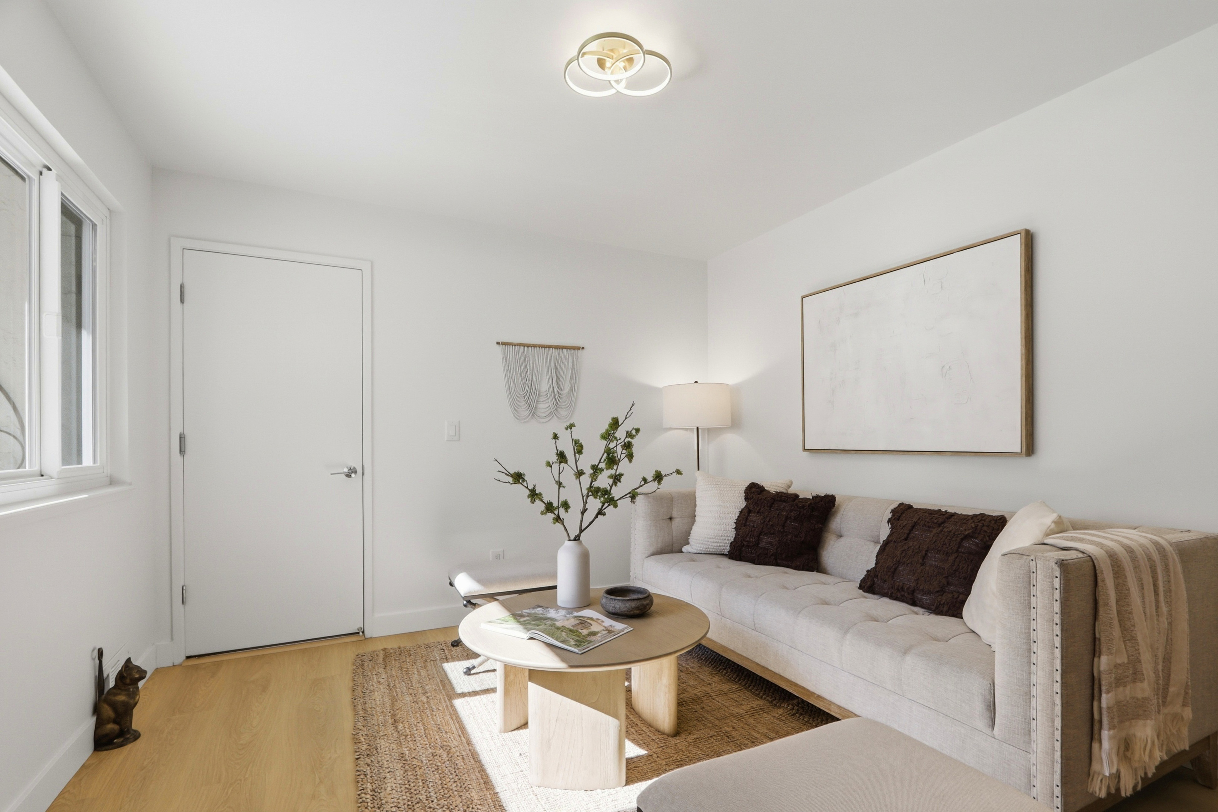 This bright and inviting living room features a neutral color palette with a tufted beige sofa, a round light-wood coffee table, and a textured jute rug. The space is accented by dark brown throw pillows, a minimalist wall hanging, and a large abstract art piece, creating a cozy and modern aesthetic. Natural light streams in from the side window, highlighting the clean lines and warm wood flooring.