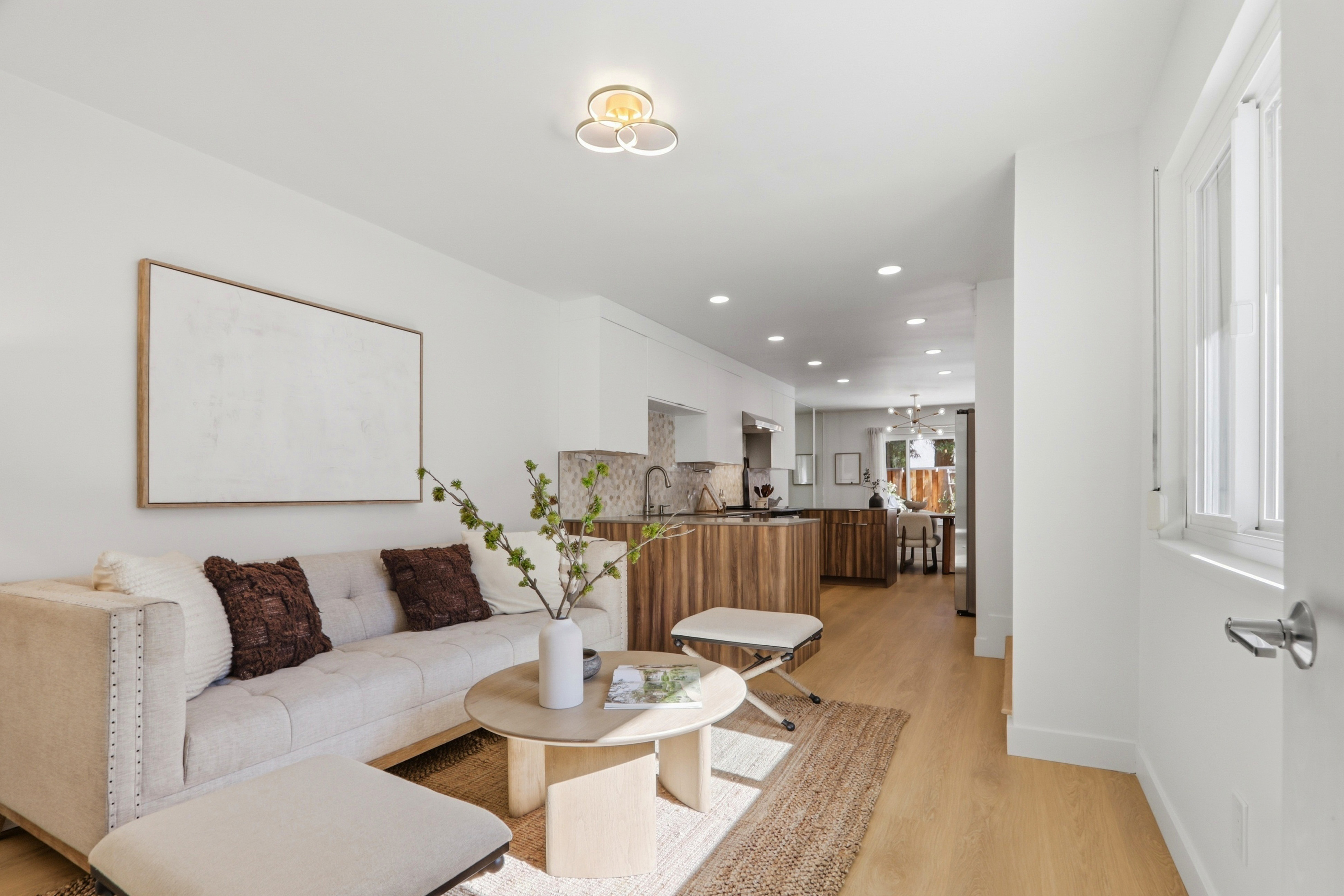 This bright and airy open-concept living space features a neutral-toned sofa with textured throw pillows, a round wooden coffee table, and a natural fiber rug. The room flows seamlessly into a modern kitchen with wood-grain cabinetry and a tiled backsplash, creating a cohesive and inviting atmosphere. The perspective is a wide-angle shot from the living area looking toward the kitchen and dining space, emphasizing the home's spacious and contemporary design.