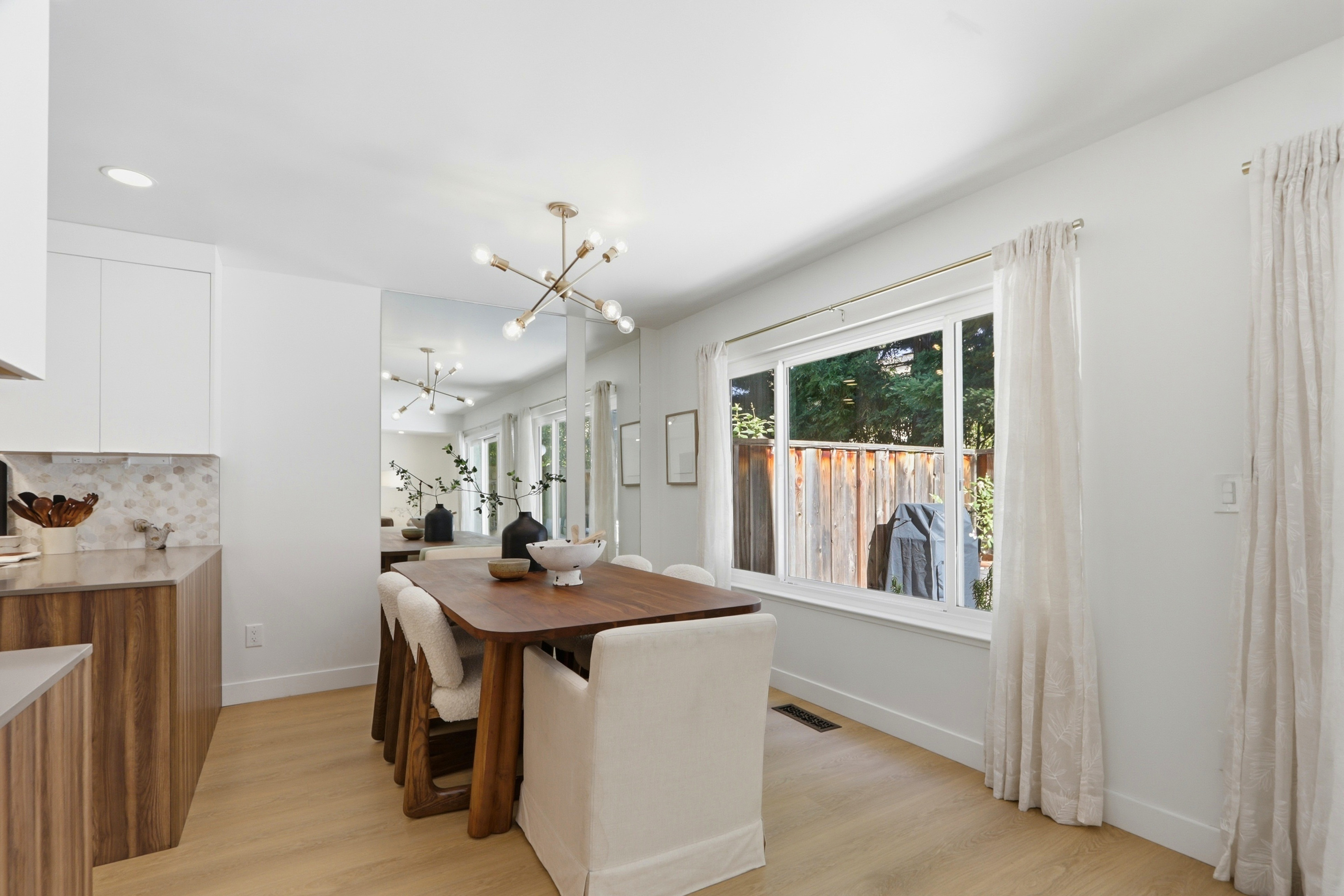 This bright and modern dining room features a warm wooden table paired with a mix of upholstered and textured chairs, set against a large wall mirror that enhances the sense of space. A contemporary sputnik-style chandelier hangs above the table, while a large window with floor-to-ceiling curtains allows for ample natural light. The room flows seamlessly into an adjacent kitchen area, creating an open and inviting atmosphere perfect for entertaining.