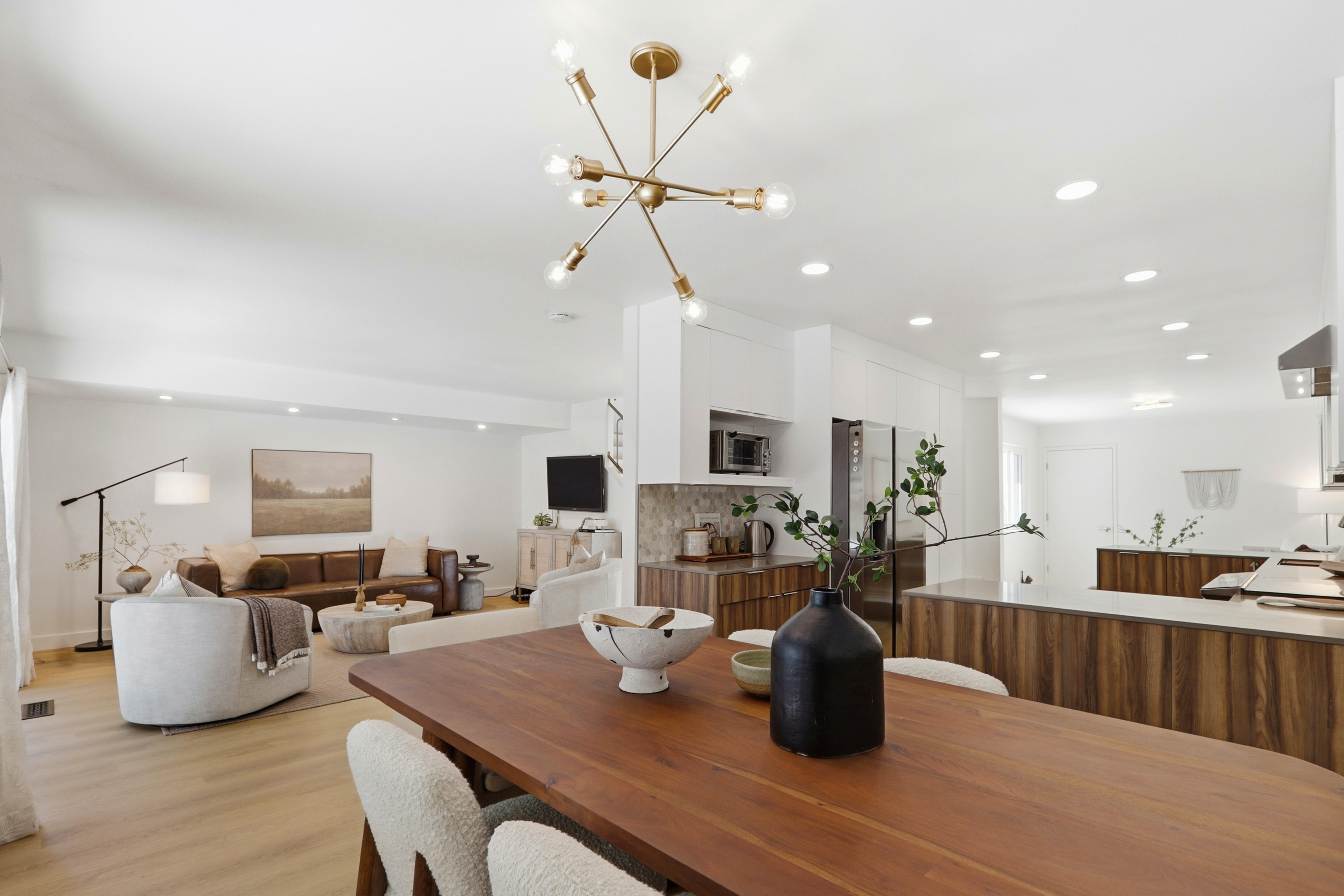 This open-concept living space seamlessly blends a modern dining area, kitchen, and living room into one cohesive, light-filled environment. The room features warm wood tones, contemporary furniture with textured upholstery, and a striking sputnik-style chandelier that serves as a focal point. The clean lines, neutral color palette, and minimalist decor create an inviting and sophisticated atmosphere perfect for modern living.