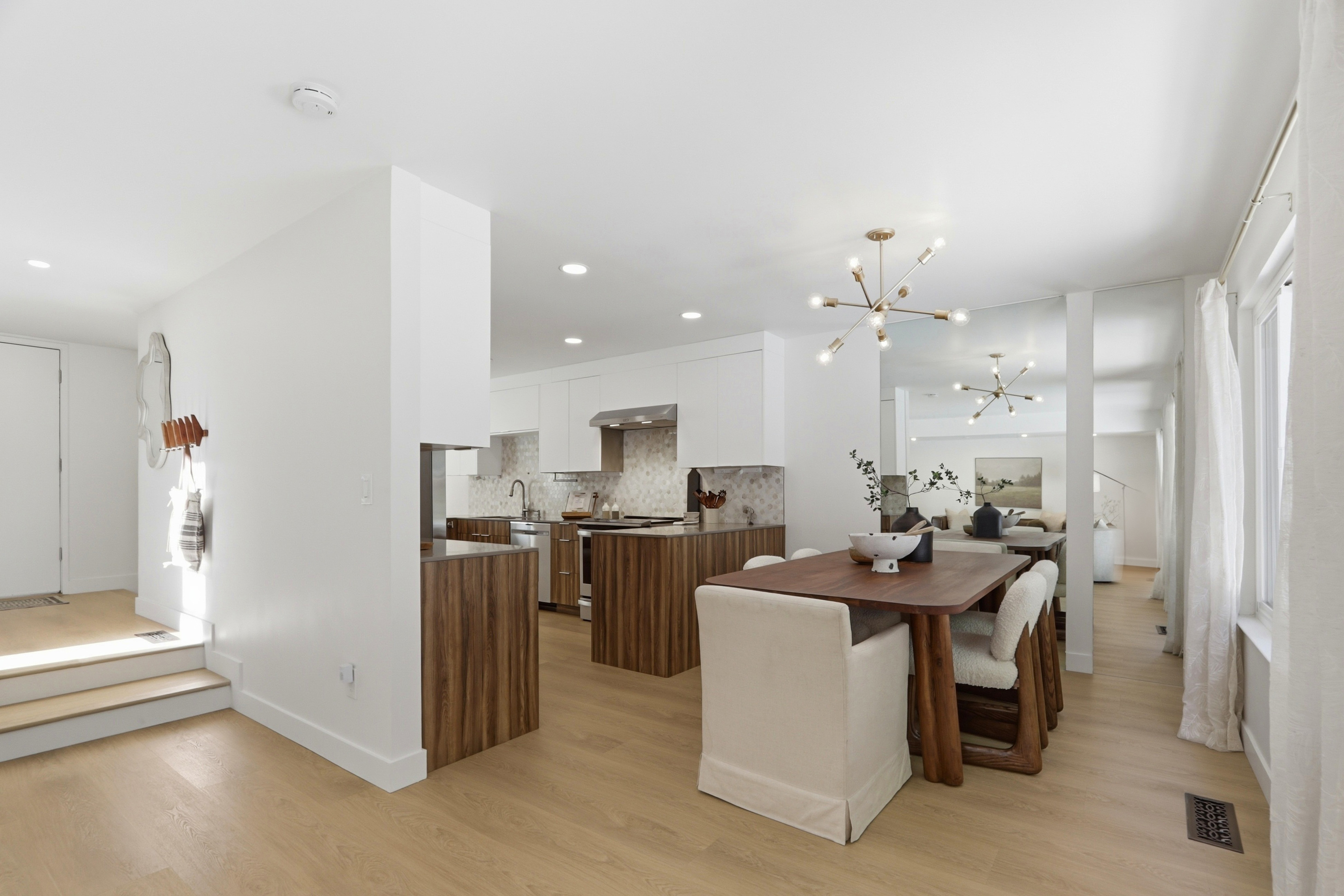 This open-concept space seamlessly blends a modern kitchen with a dining area, featuring a striking contrast between white upper cabinetry and rich, wood-grain lower cabinets and island. The kitchen is equipped with stainless steel appliances and a decorative mosaic backsplash, while the dining area is anchored by a warm wooden table and upholstered chairs under a contemporary sputnik chandelier. The light wood flooring and neutral color palette create an airy, inviting atmosphere that flows into the adjacent living space.