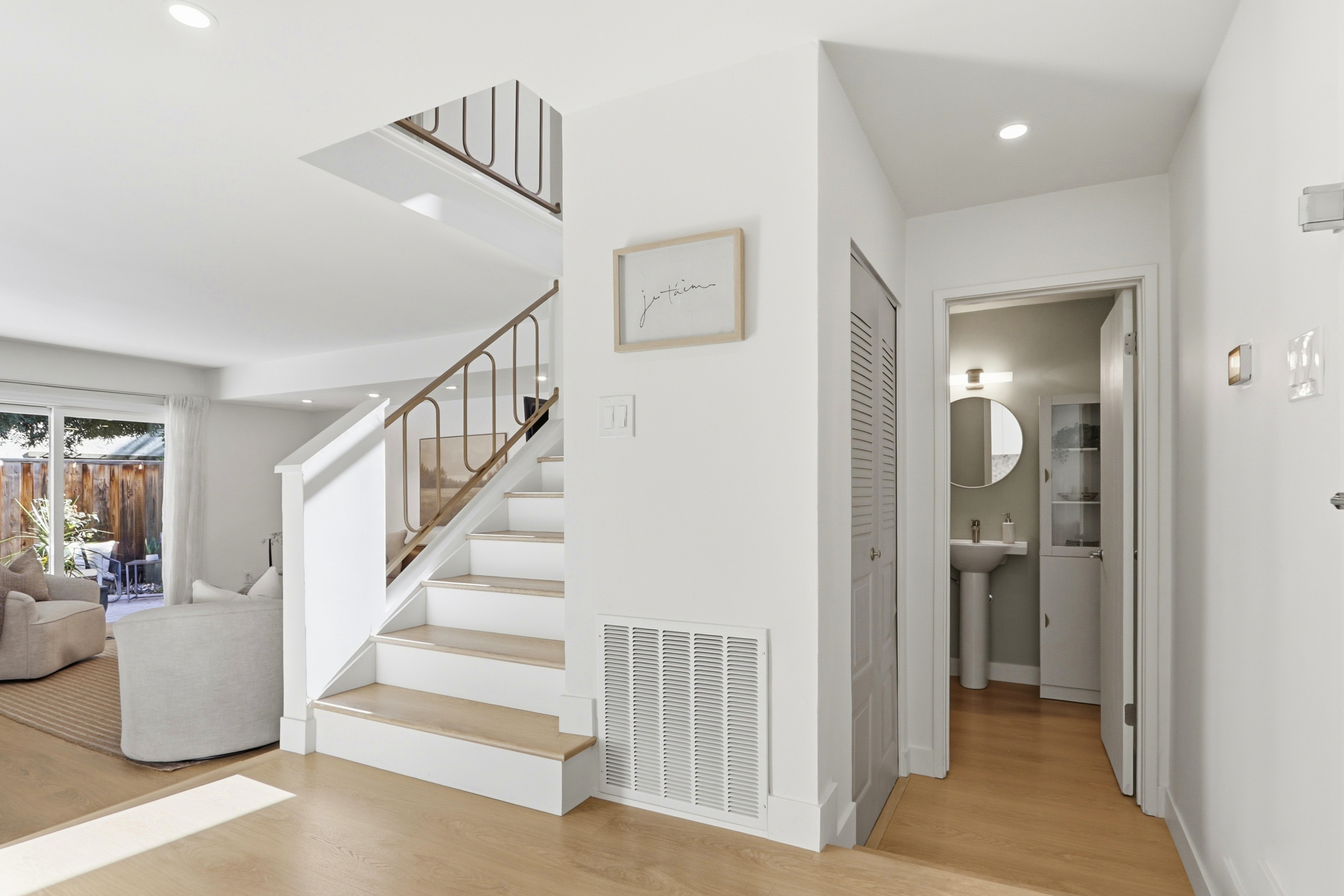 This bright and airy interior space features a staircase with light wood treads and a modern, minimalist metal railing. To the right, a doorway leads into a clean, neutral-toned bathroom, while the left side opens into a living area with a view of a patio. The home is characterized by a crisp white color palette, light hardwood flooring, and recessed lighting, creating a welcoming and contemporary atmosphere.